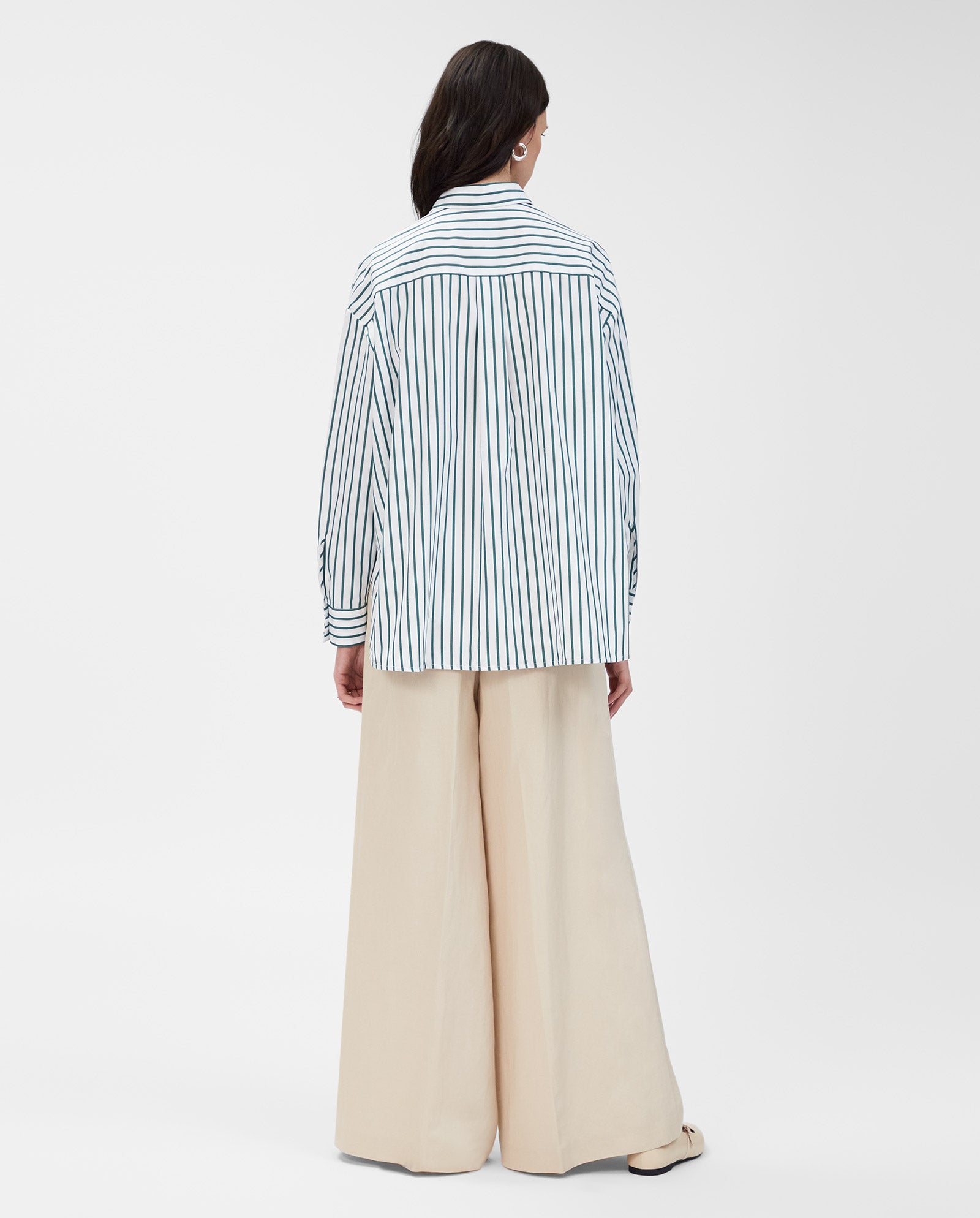 A woman faces away, wearing the IVY OAK BETHANY LILLY blue-striped cotton blouse with a boxy cut and wide-leg beige pants against a plain white background.