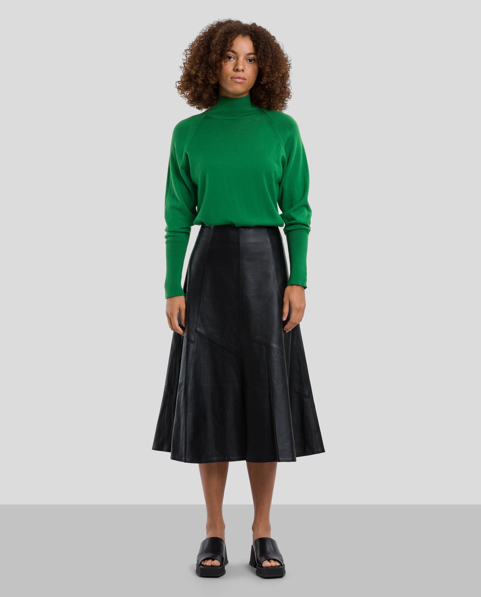 A woman wears the IVY OAK LAUREEN Leather Skirt in sleek black, paired with a green turtleneck sweater, standing against a plain gray background.