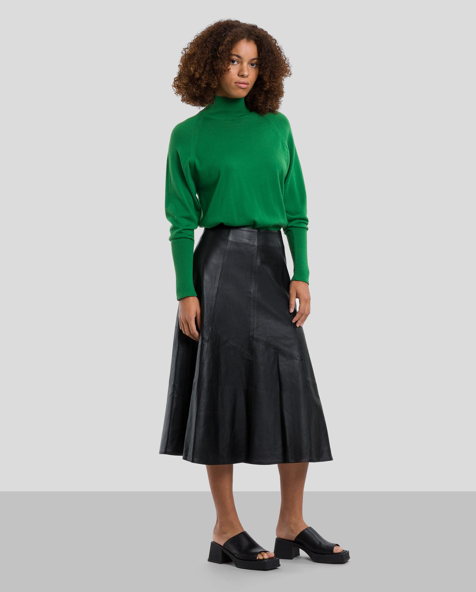 A woman stands on gray wearing a green turtleneck and the IVY OAK LAUREEN Leather Skirt, a black chrome-free leather midi skirt, paired with black open-toe sandals.