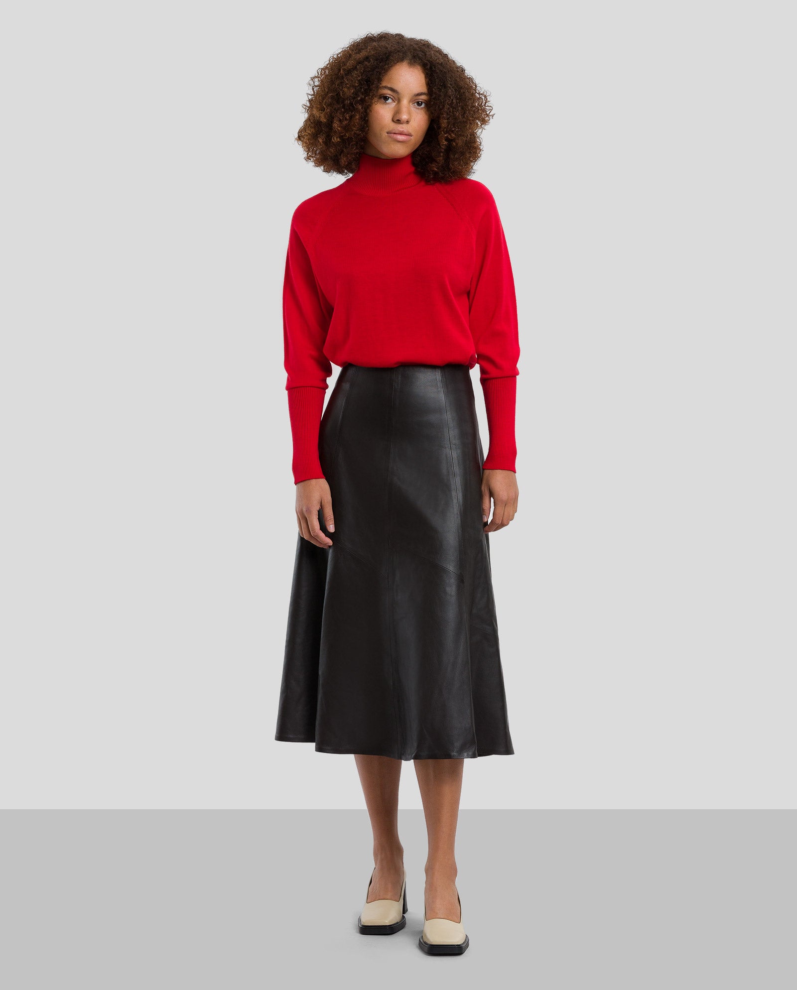 A woman wears the IVY OAK LAUREEN Leather Skirt in black, paired with a red turtleneck sweater and cream-colored shoes.