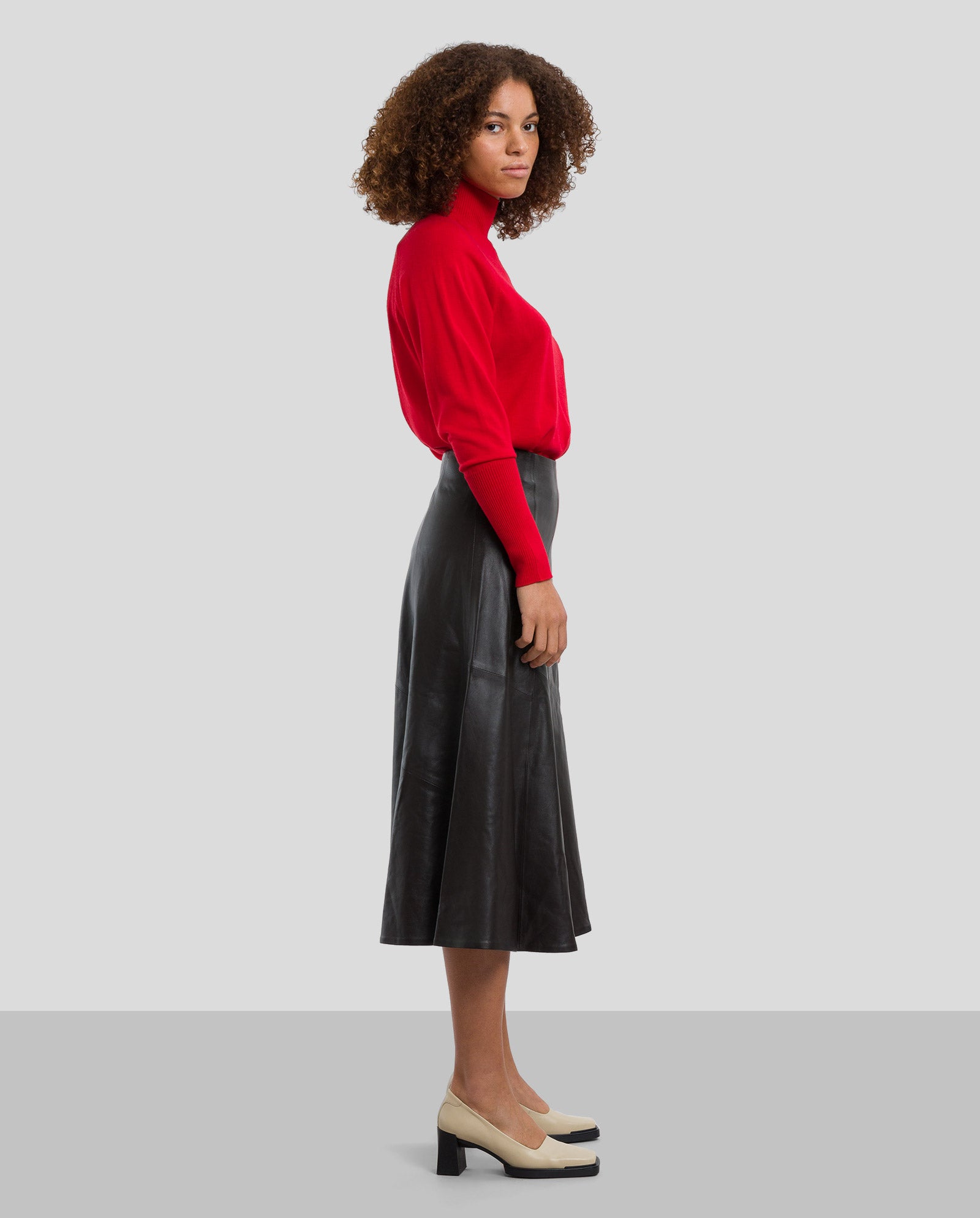 A woman in a red turtleneck and beige heels stands in profile against a gray background, wearing the IVY OAK LAUREEN Leather Skirt crafted from A-shaped chrome-free tanned leather.
