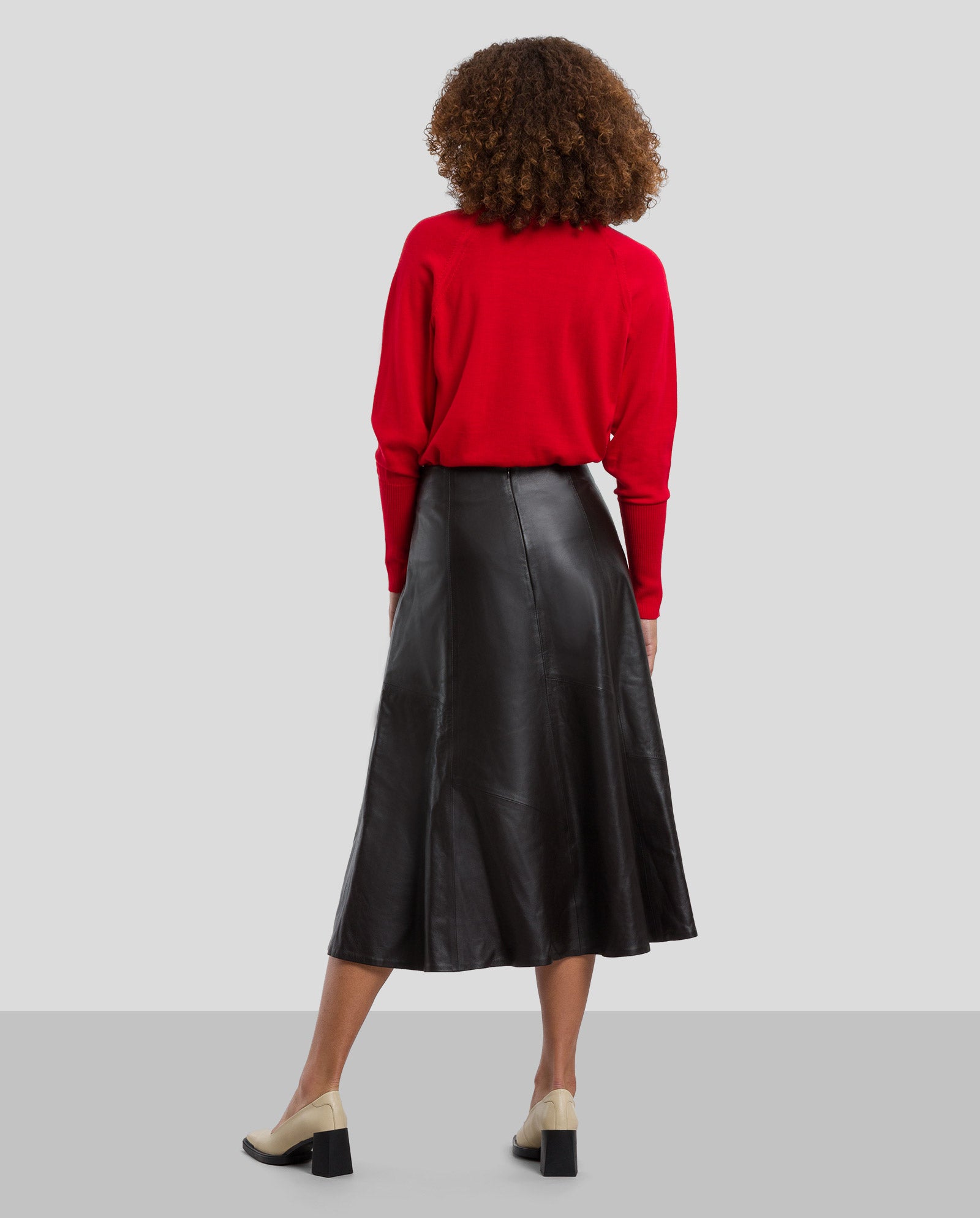 A woman faces away, wearing a red sweater and the IVY OAK LAUREEN Leather Skirt in A-line, chrome-free tanned black leather, paired with beige heels against a plain background.