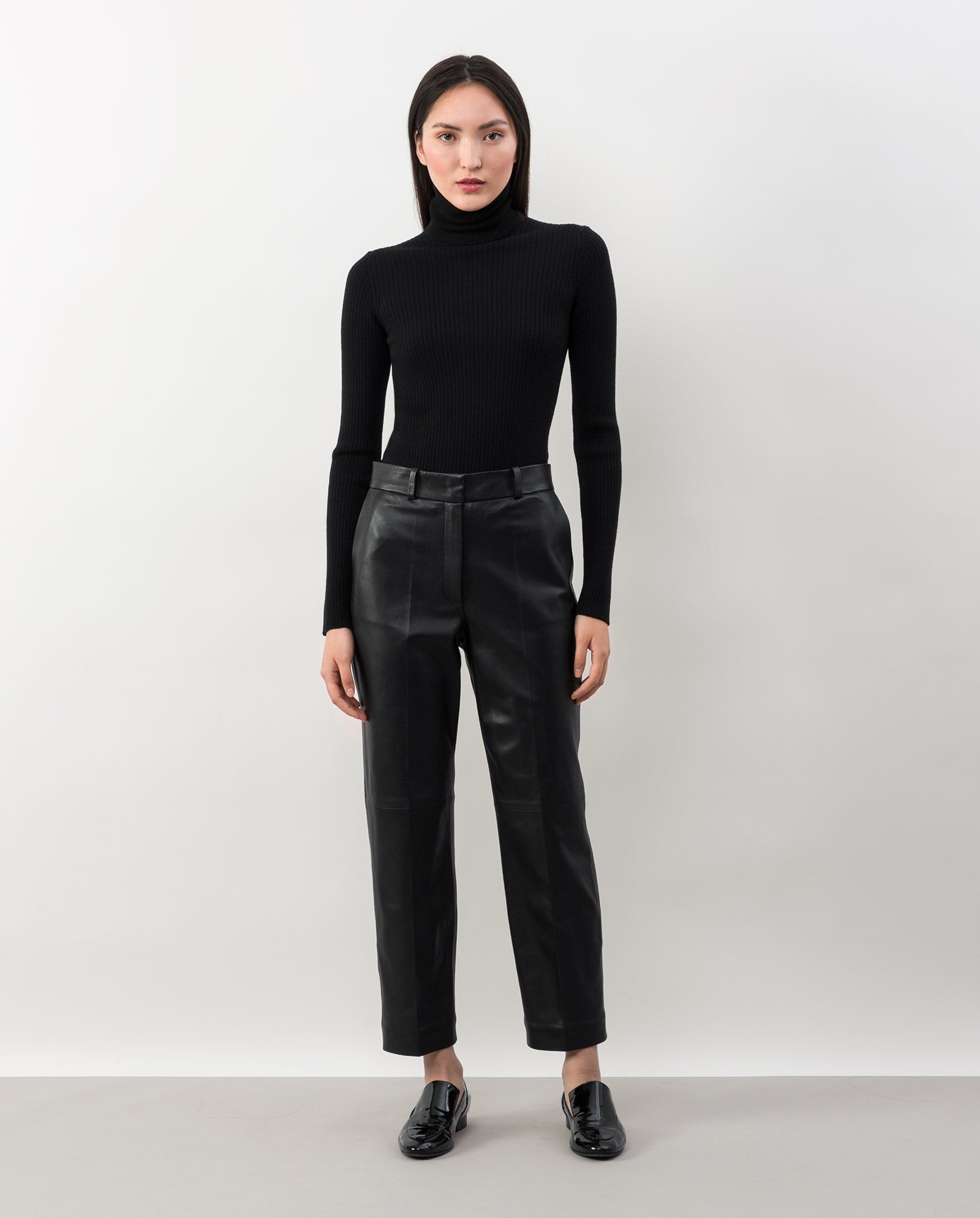 A woman stands against a plain background wearing black loafers, a black turtleneck, and IVY OAK’s LAURA Leather Trousers—slim fit and crafted from chrome-free leather.