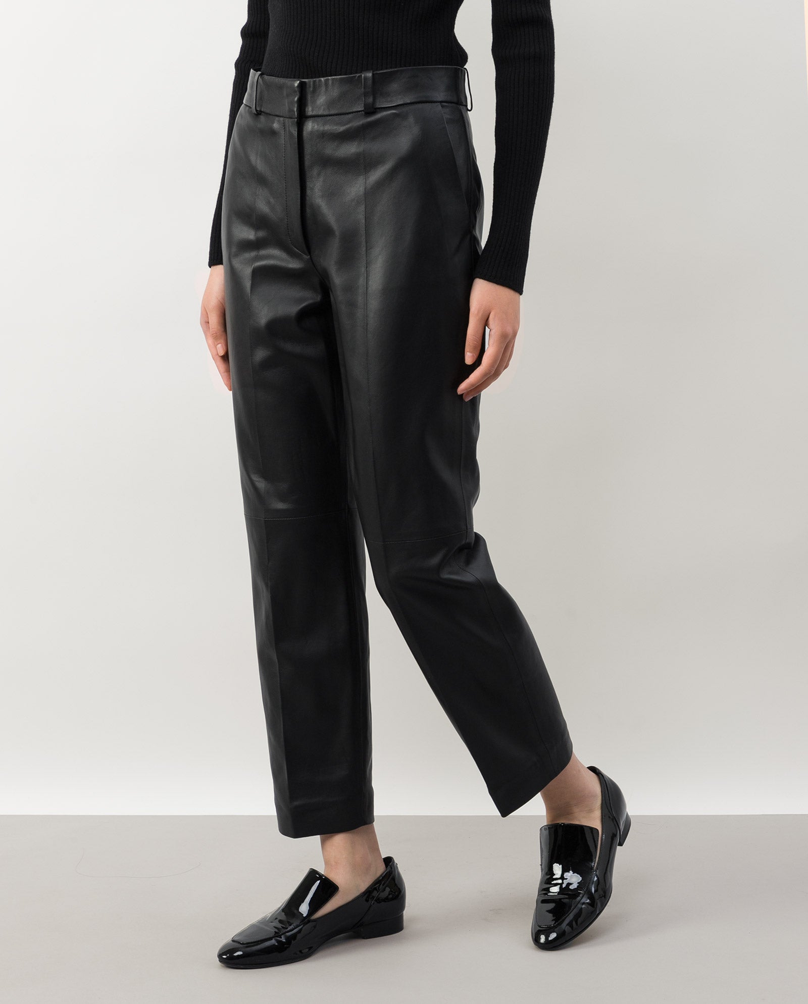 A person wears IVY OAK’s LAURA Leather Trousers in black, paired with black shiny shoes and a black ribbed sweater, standing against a plain background.