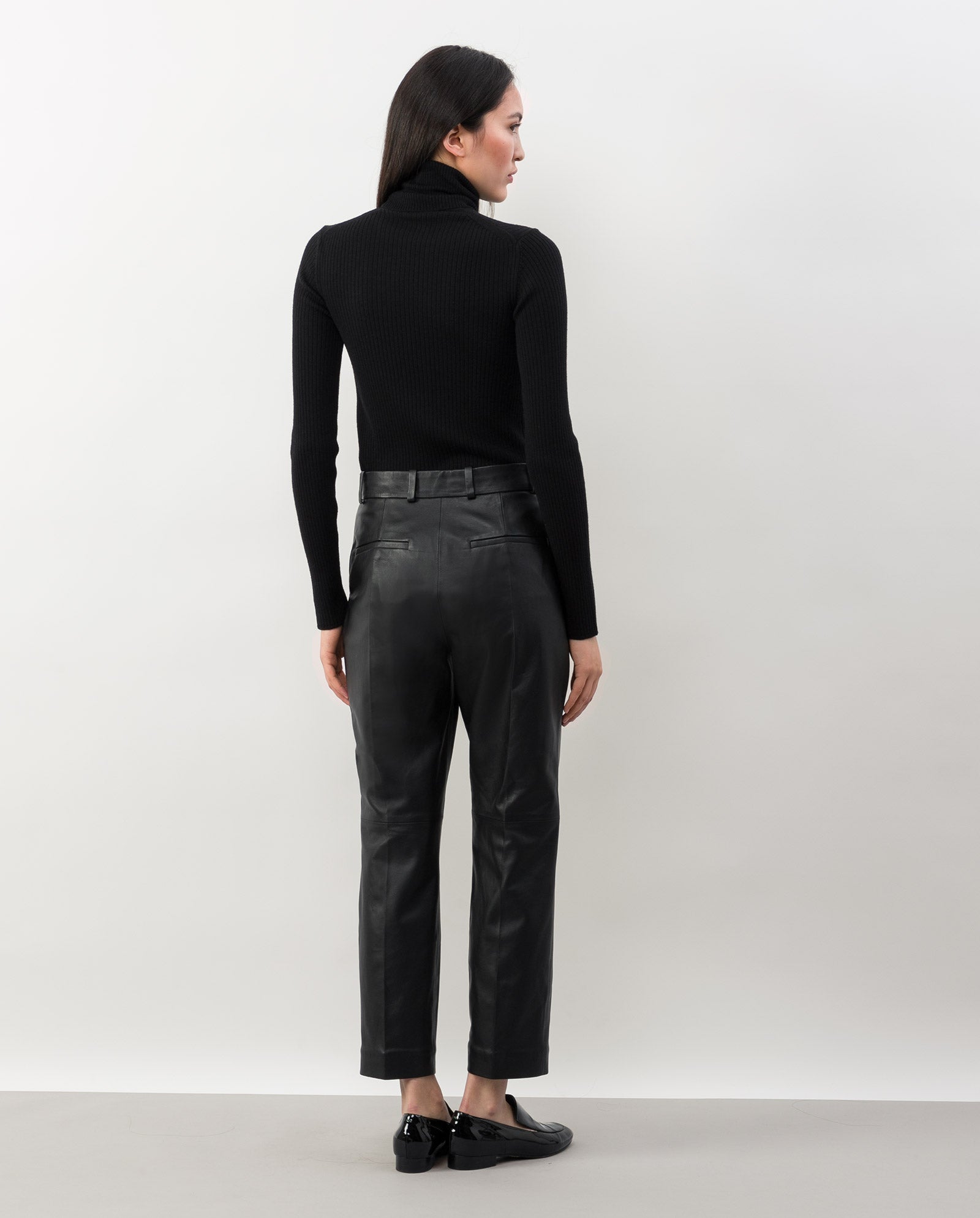 A woman faces away from the camera in a black turtleneck, IVY OAK LAURA leather trousers, and black shoes, set against a white background.