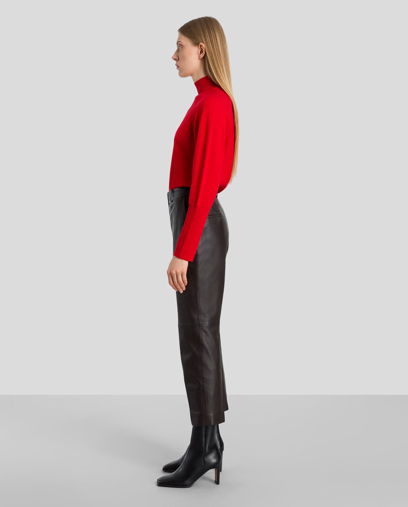 A woman in a red turtleneck and black heeled boots models IVY OAKs LAURA Leather Trousers, standing in profile against a gray background.