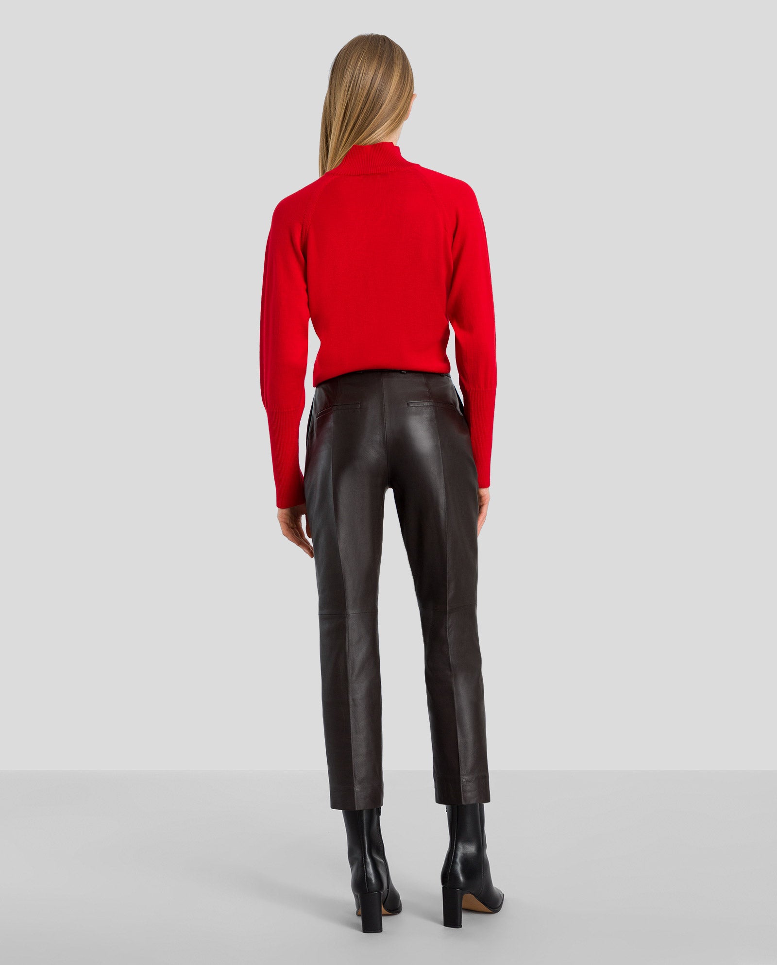 A woman faces away, wearing a red long-sleeve top, IVY OAK LAURA Leather Trousers in black high waist, and black heeled boots.