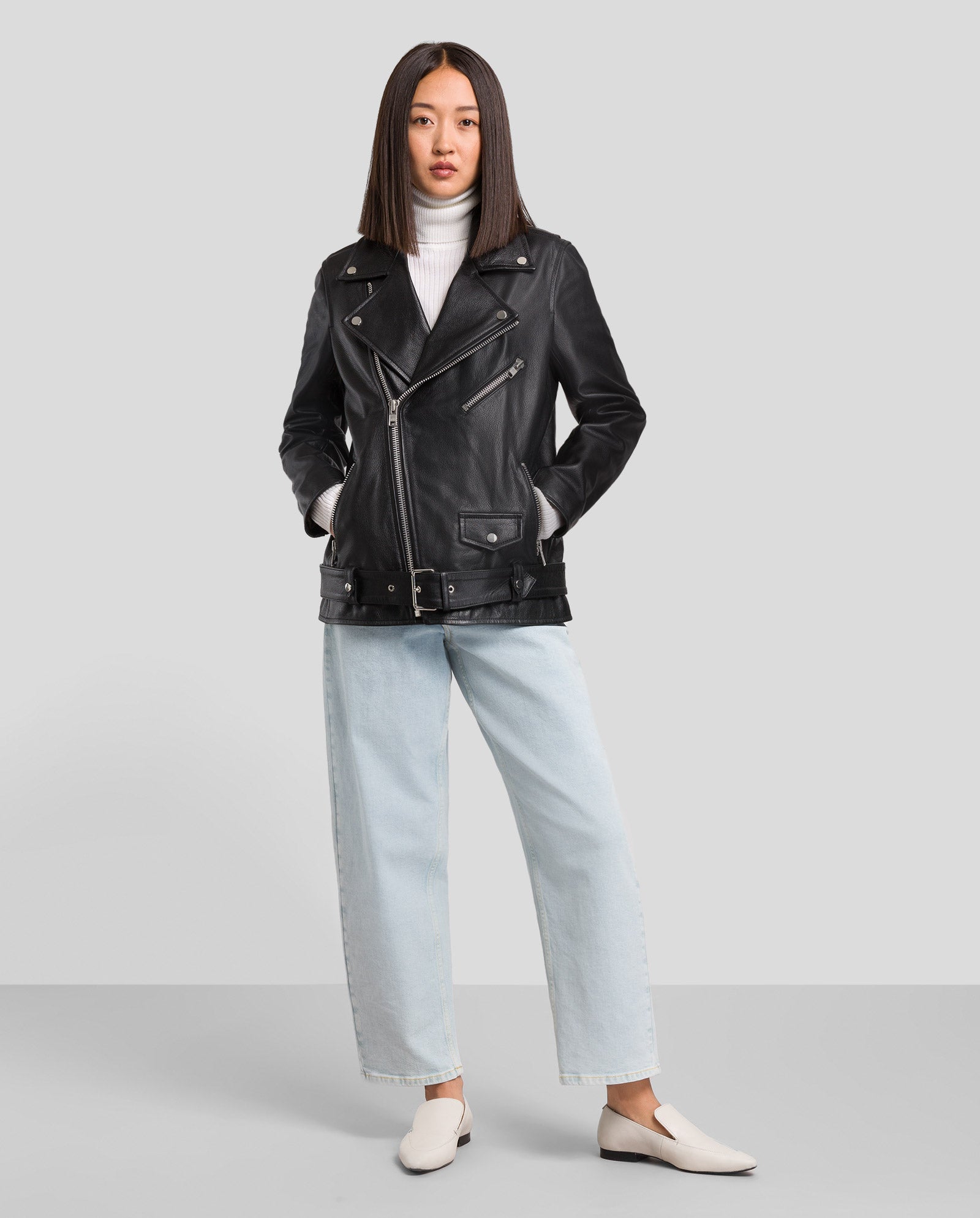 Eine Frau trägt die locker geschnittene LENNY Lederjacke von IVY OAK in Chrom-freiem Leder, kombiniert mit weißem Rollkragen, hellblauen Jeans und weißen Schuhen vor grauem Hintergrund.