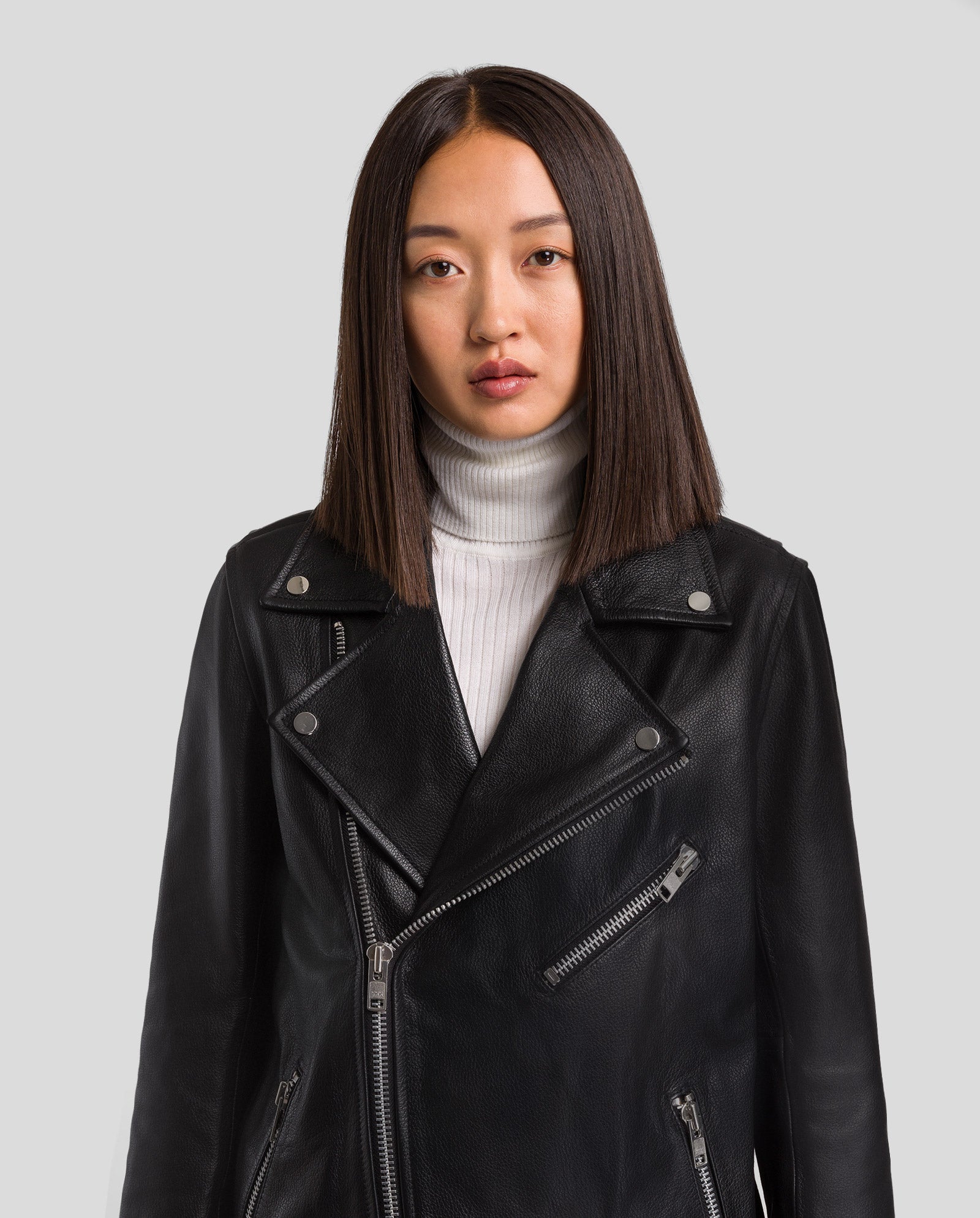 Eine Frau trägt die LENNY Leather Jacket von IVY OAK in lockerer Passform und weißem Turtleneck vor schlichtem, hellgrauem Hintergrund. Die Jacke ist chromfrei gefertigt.