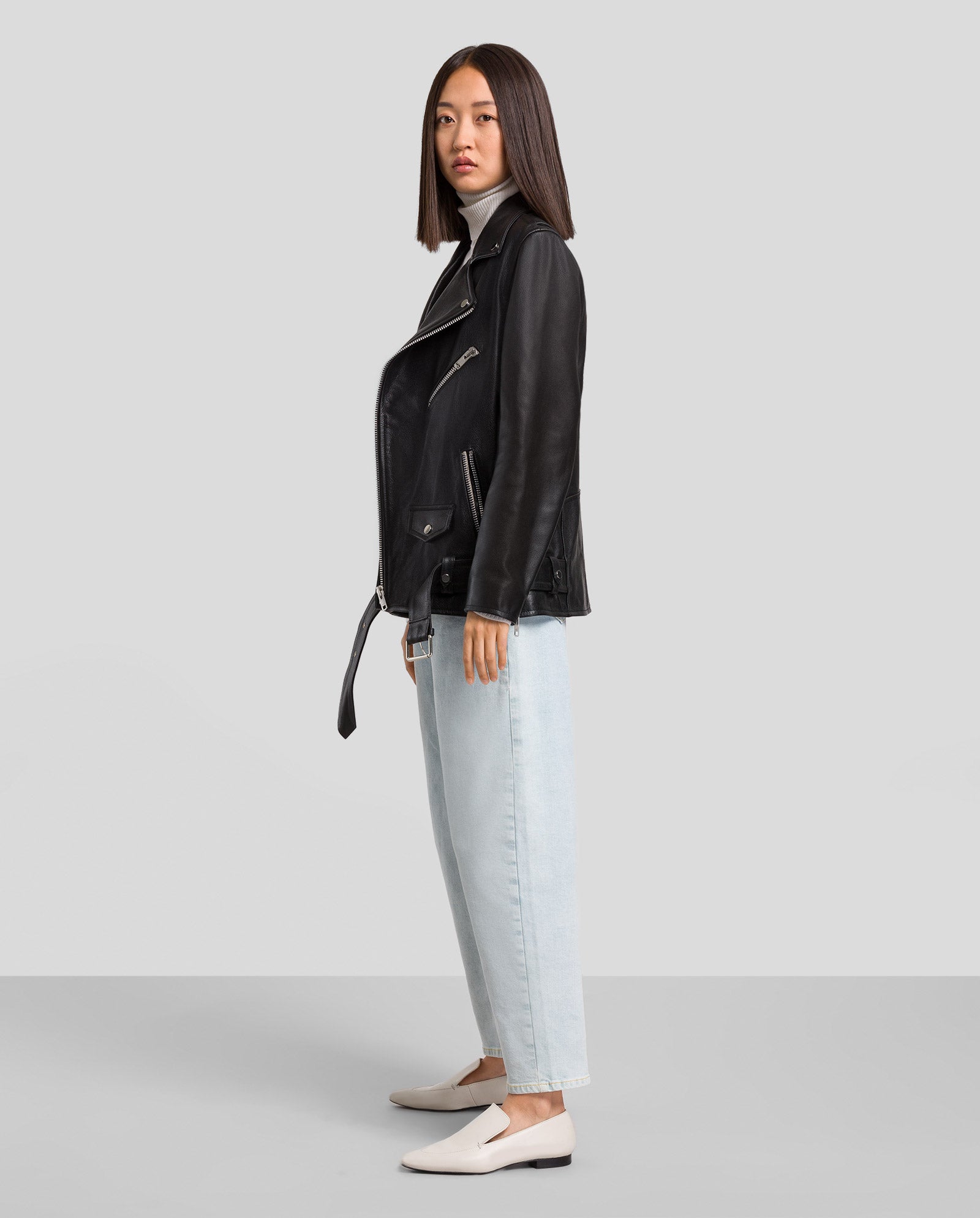 Frau trägt die LENNY Leather Jacket von IVY OAK mit lockerer Passform, kombiniert mit hellen Jeans und weißen Schuhen, und steht seitlich vor schlichtem Hintergrund.
