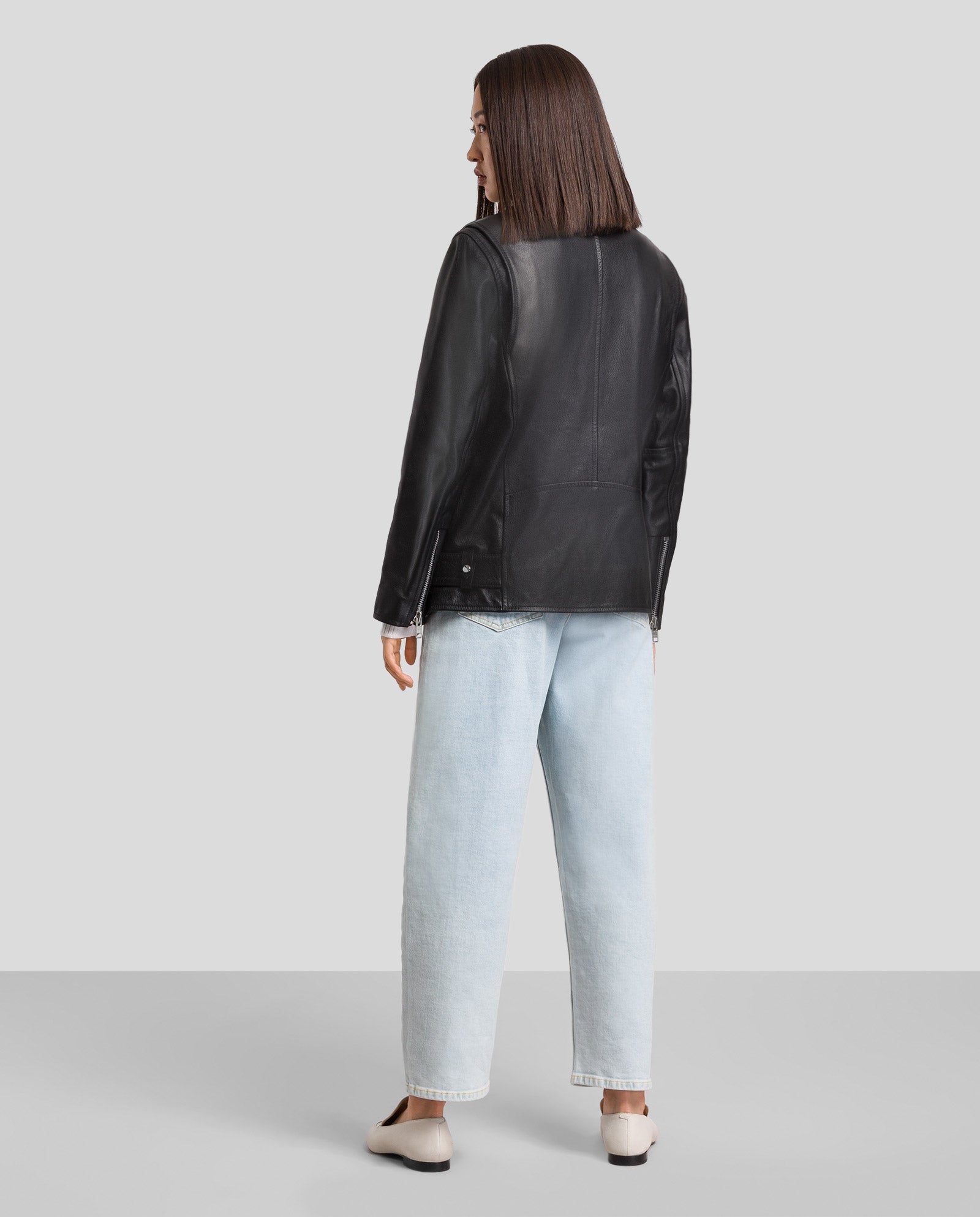 Person trägt die LENNY Leather Jacket von IVY OAK aus chromfreiem Leder und hellblaue Jeans mit lockerer Passform, während sie mit dem Rücken zur Kamera steht.