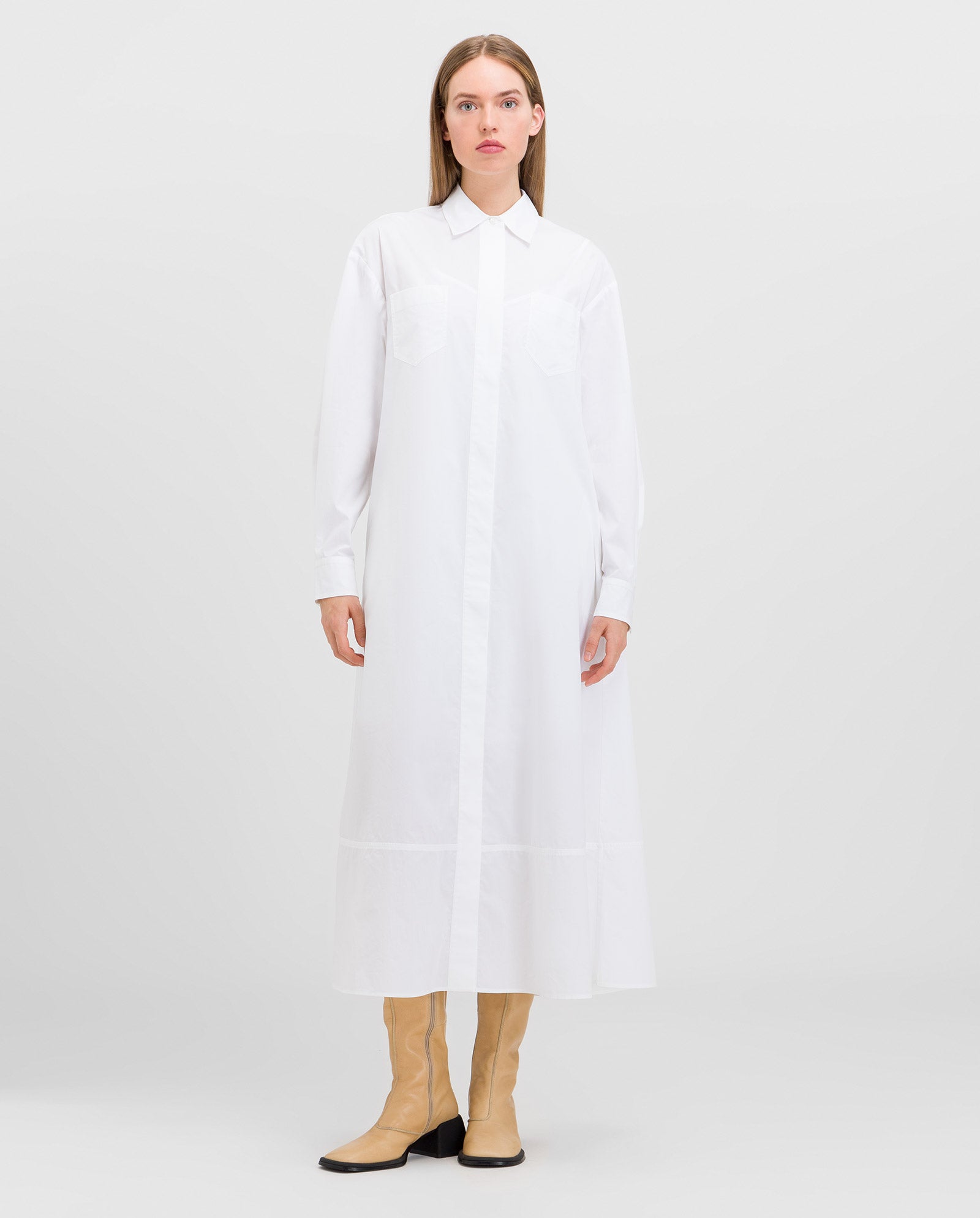 A woman stands against a plain light background wearing the IVY OAK DINA ANN Dress—an ankle-length, white flared shirt dress crafted from organic fabric—styled with tan knee-high boots.
