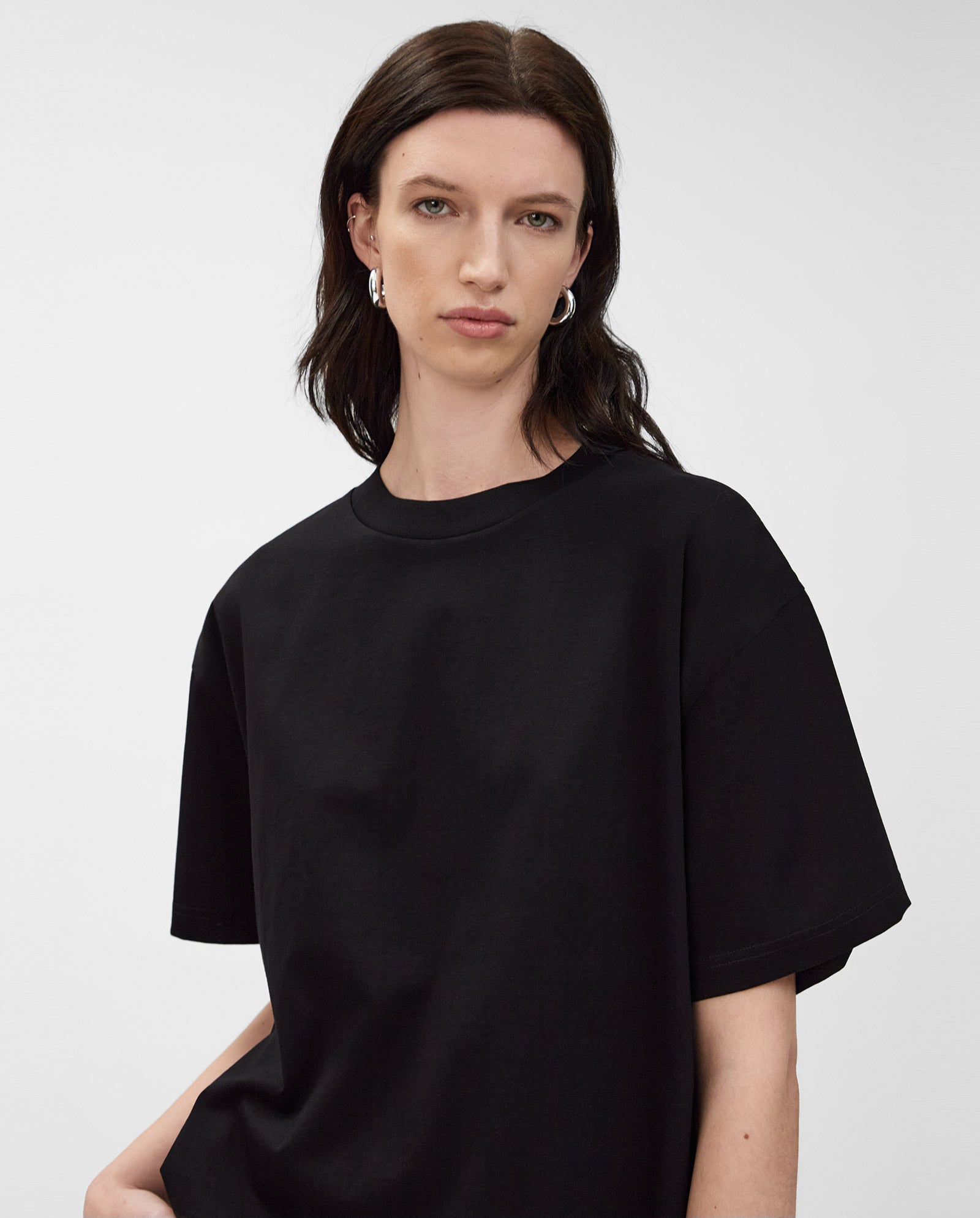 A woman with long dark hair wears the KYLA T-Shirt by IVY OAK and silver hoop earrings, standing against a plain background.