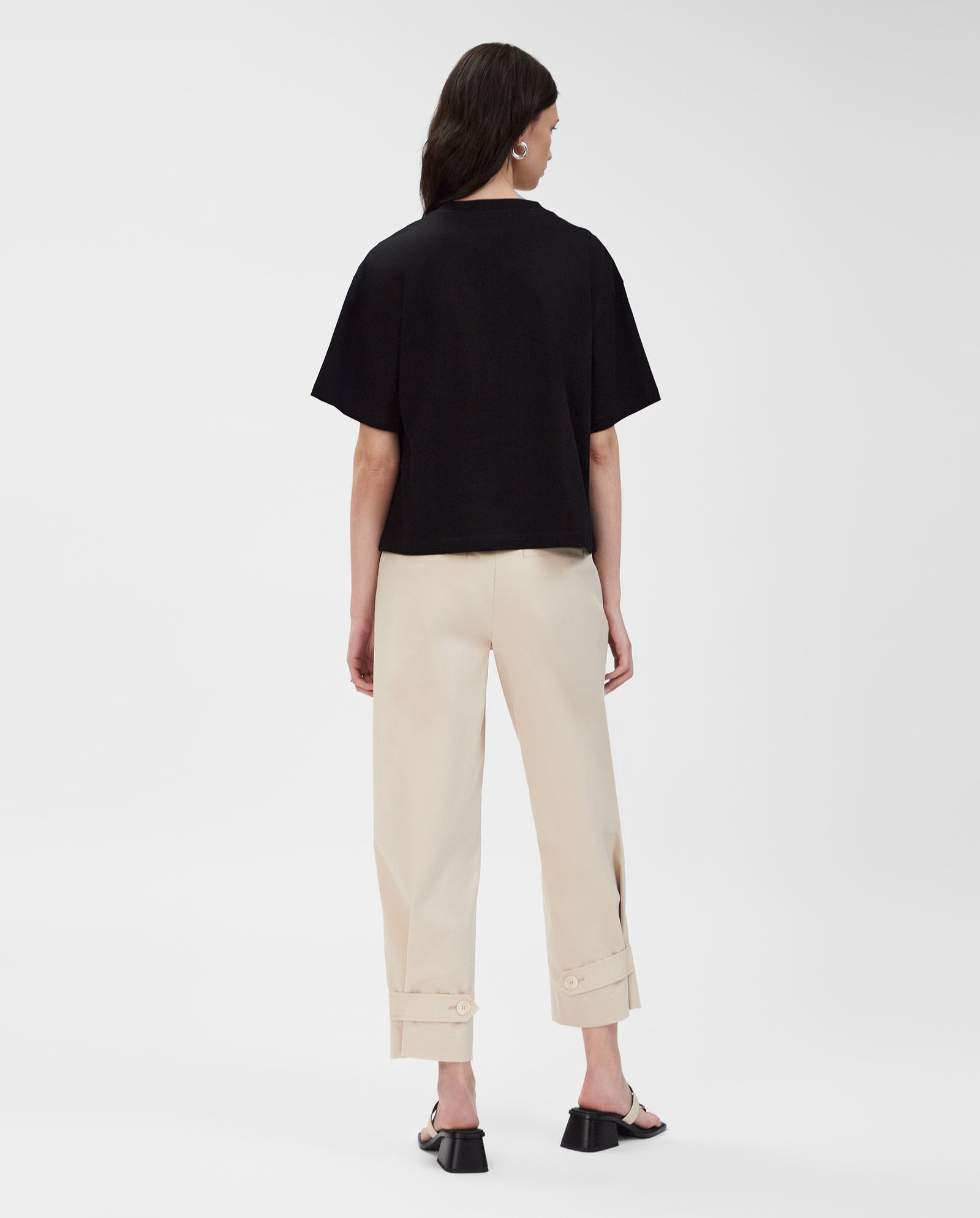 A woman stands with her back to the camera, wearing the IVY OAK KYLA T-Shirt in black, paired with beige pants and black sandals.