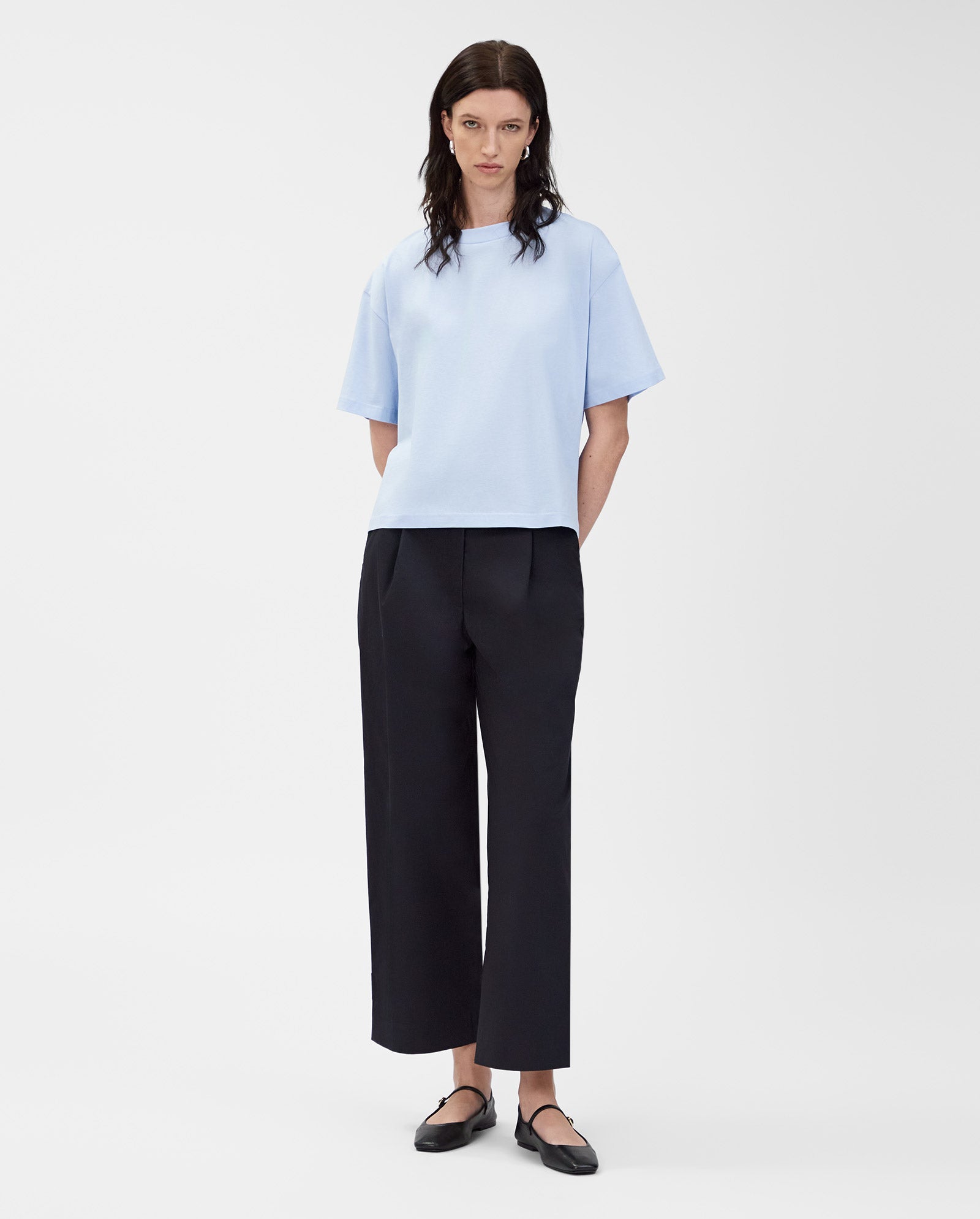 A person wears an IVY OAK KYLA T-Shirt in light blue organic cotton with black wide-leg pants and black shoes, standing against a white background—a wardrobe essential.