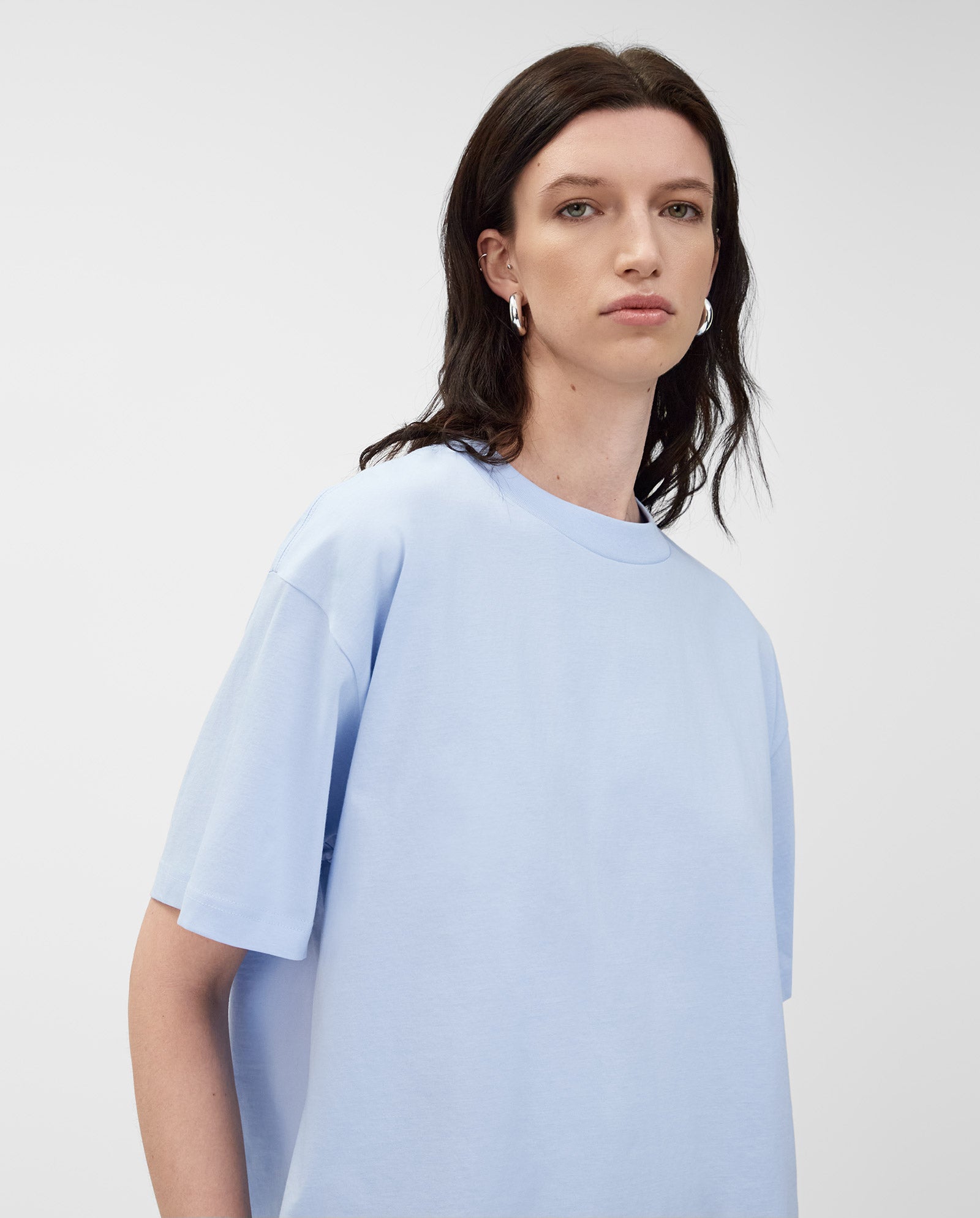 A person with long dark hair wears a light blue KYLA T-shirt by IVY OAK, crafted from organic cotton, and silver hoop earrings, standing against a plain background—a timeless wardrobe essential.