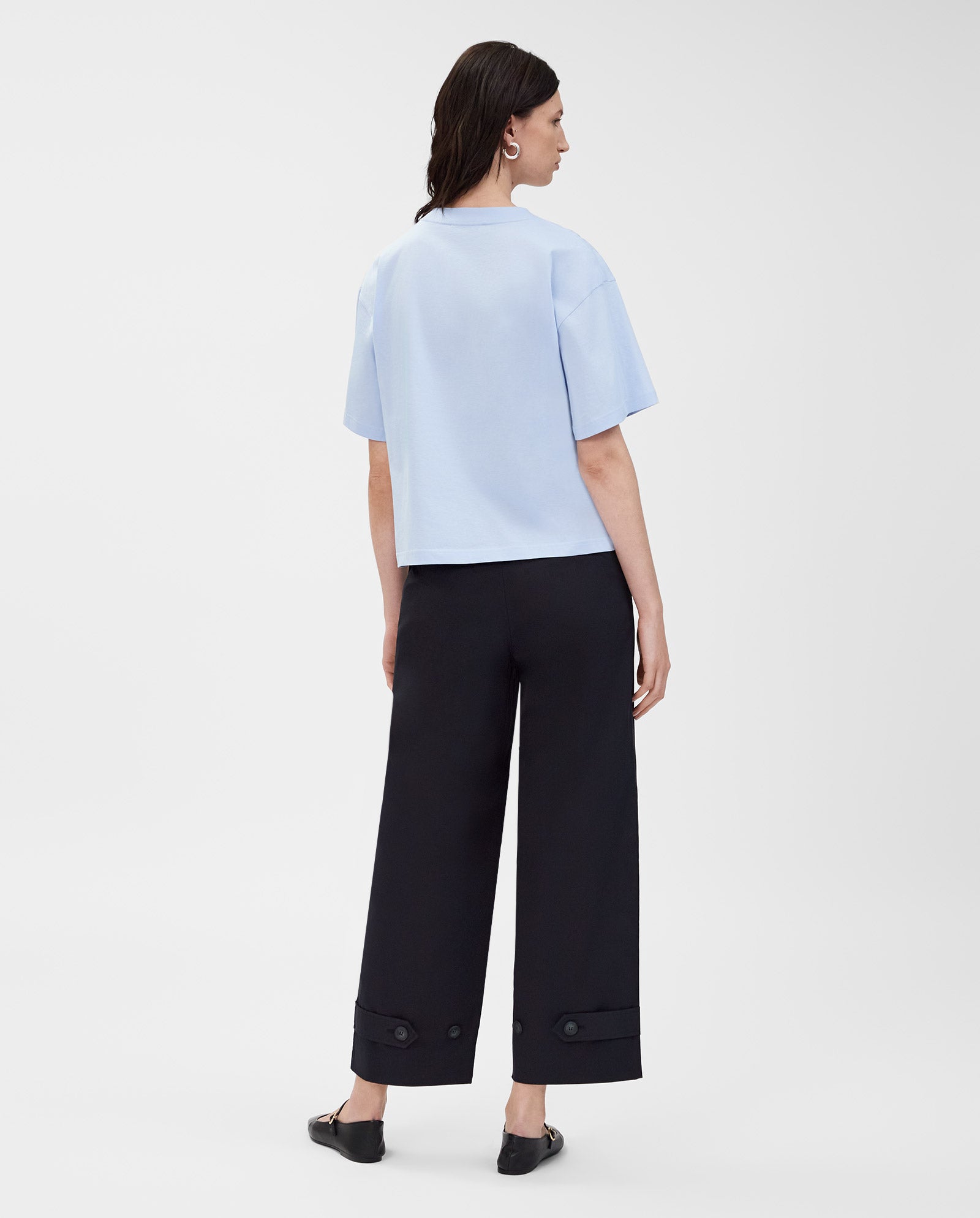 Woman standing, facing away, wears the IVY OAK KYLA T-Shirt in light blue—an organic cotton essential—paired with wide-leg black pants and black shoes.
