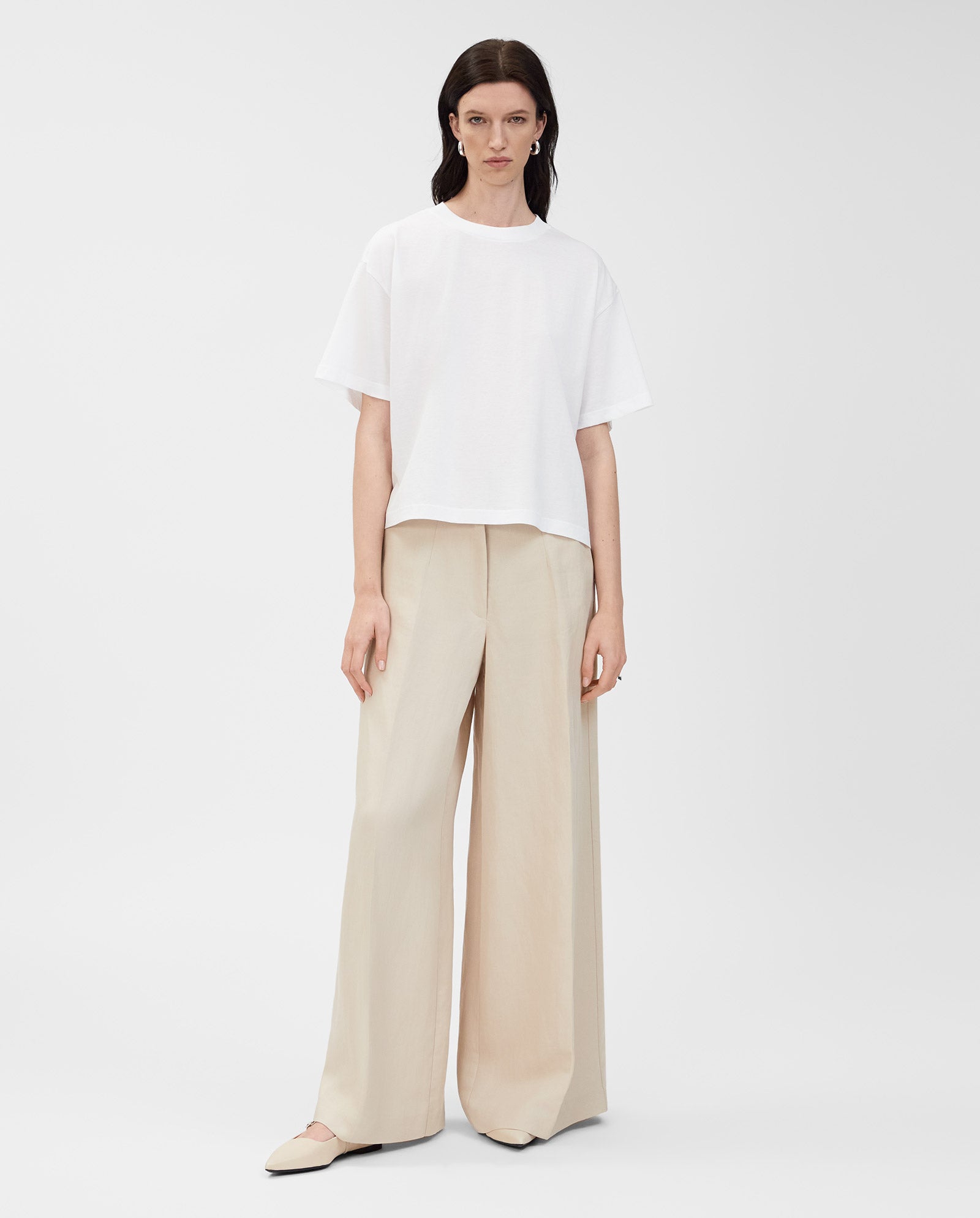 A woman wears the IVY OAK KYLA T-Shirt in organic cotton with wide beige pants and beige flats, standing against a plain white background.
