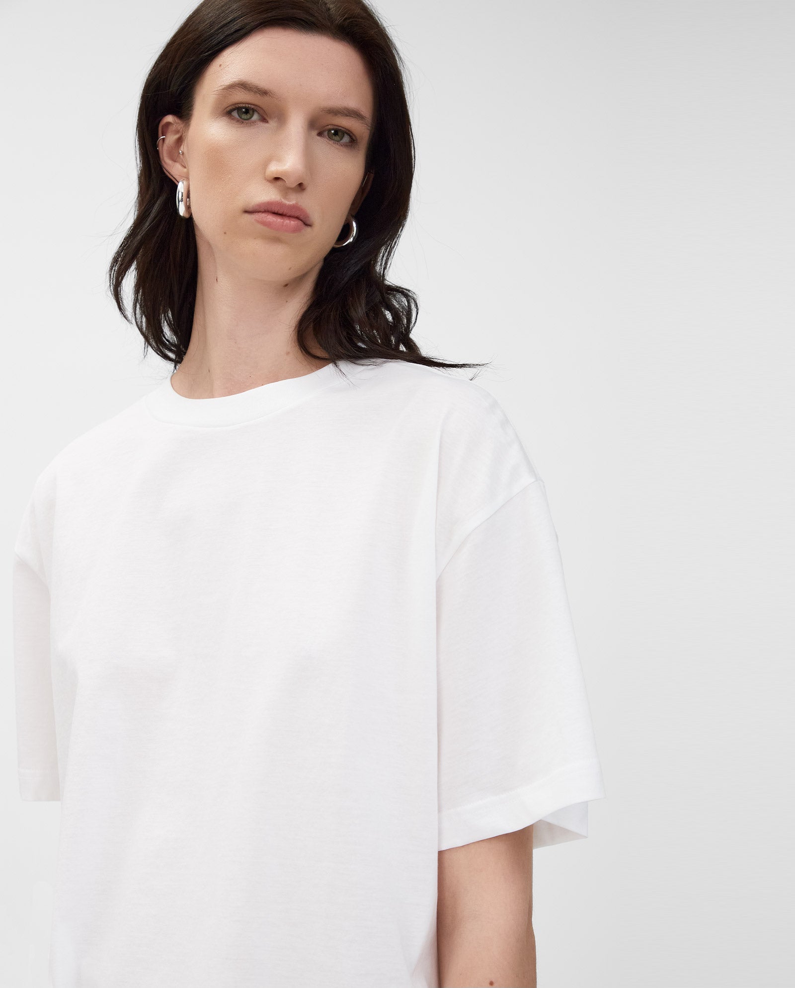 Woman with dark hair wears the KYLA T-Shirt by IVY OAK, made from organic cotton—a basic must-have—paired with silver hoop earrings, standing against a plain background.