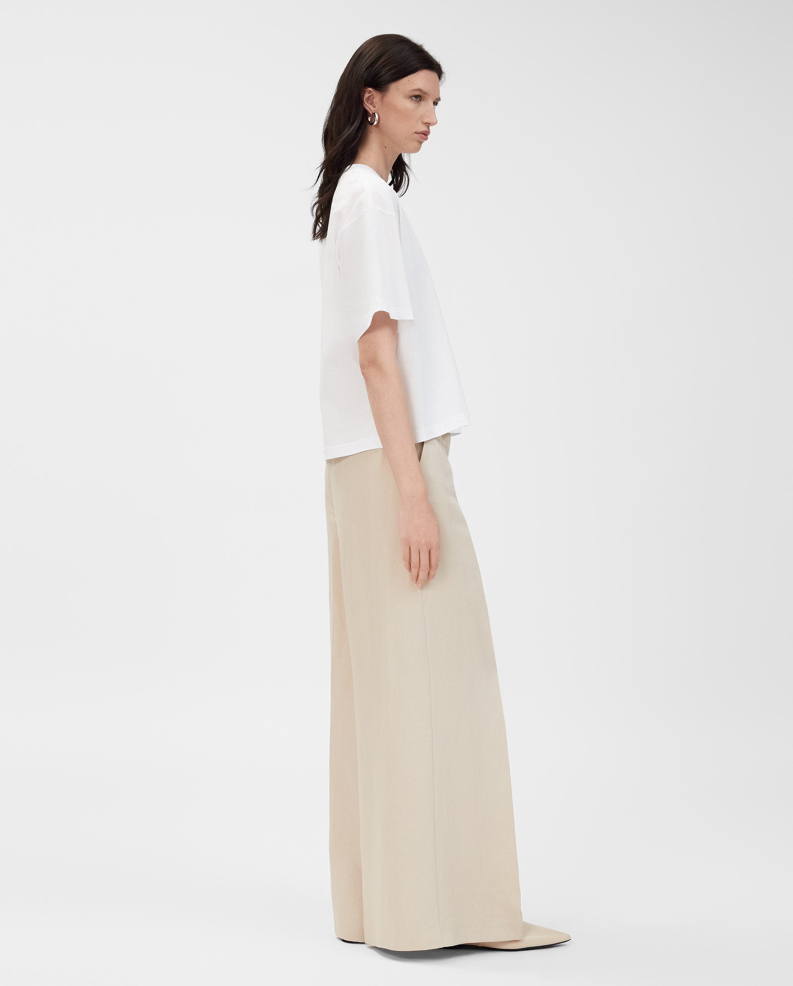 A woman wearing the IVY OAK KYLA T-Shirt, made from organic cotton, and wide-leg beige pants stands sideways against a plain white background.
