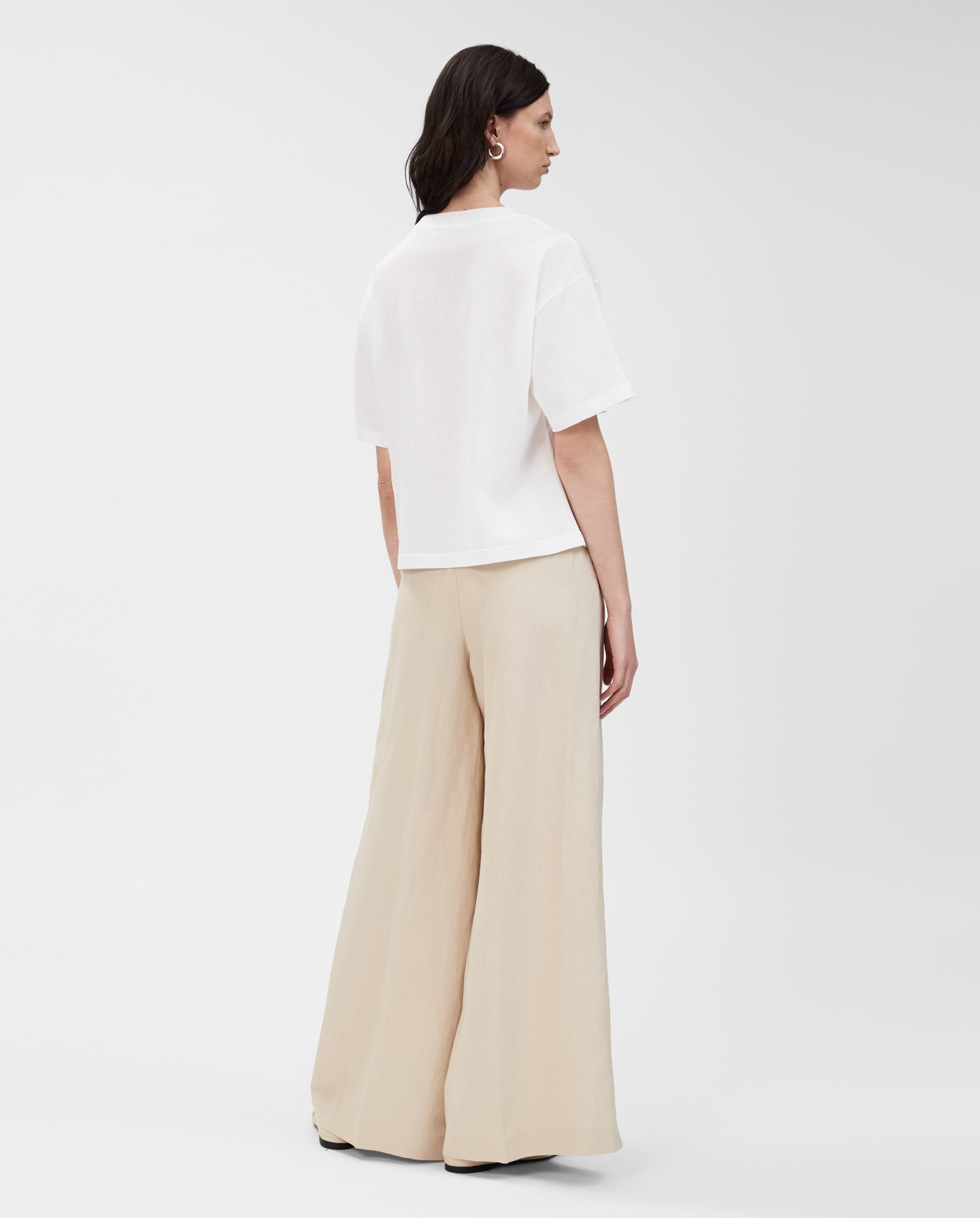 A woman stands facing away, wearing the IVY OAK KYLA T-Shirt in white—an essential piece made from organic cotton—paired with wide-leg beige pants, set against a plain background.