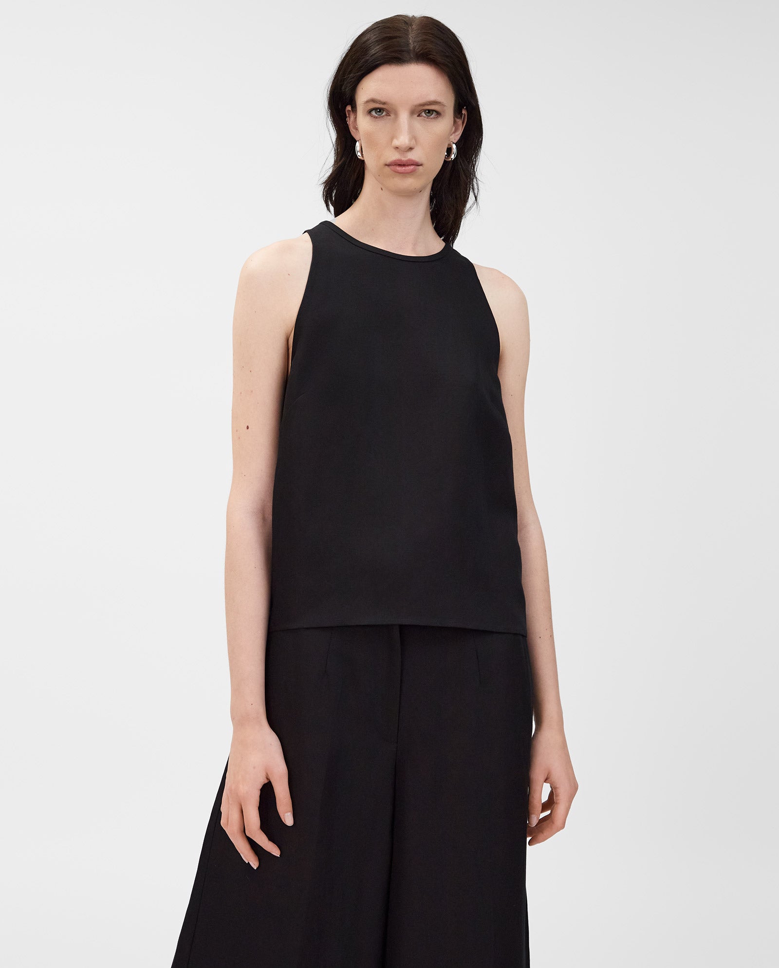 A woman stands against a plain white background, wearing the IVY OAK TAILOR SUE Top—a sleeveless American shoulder design—paired with wide-leg black pants in a linen blend.