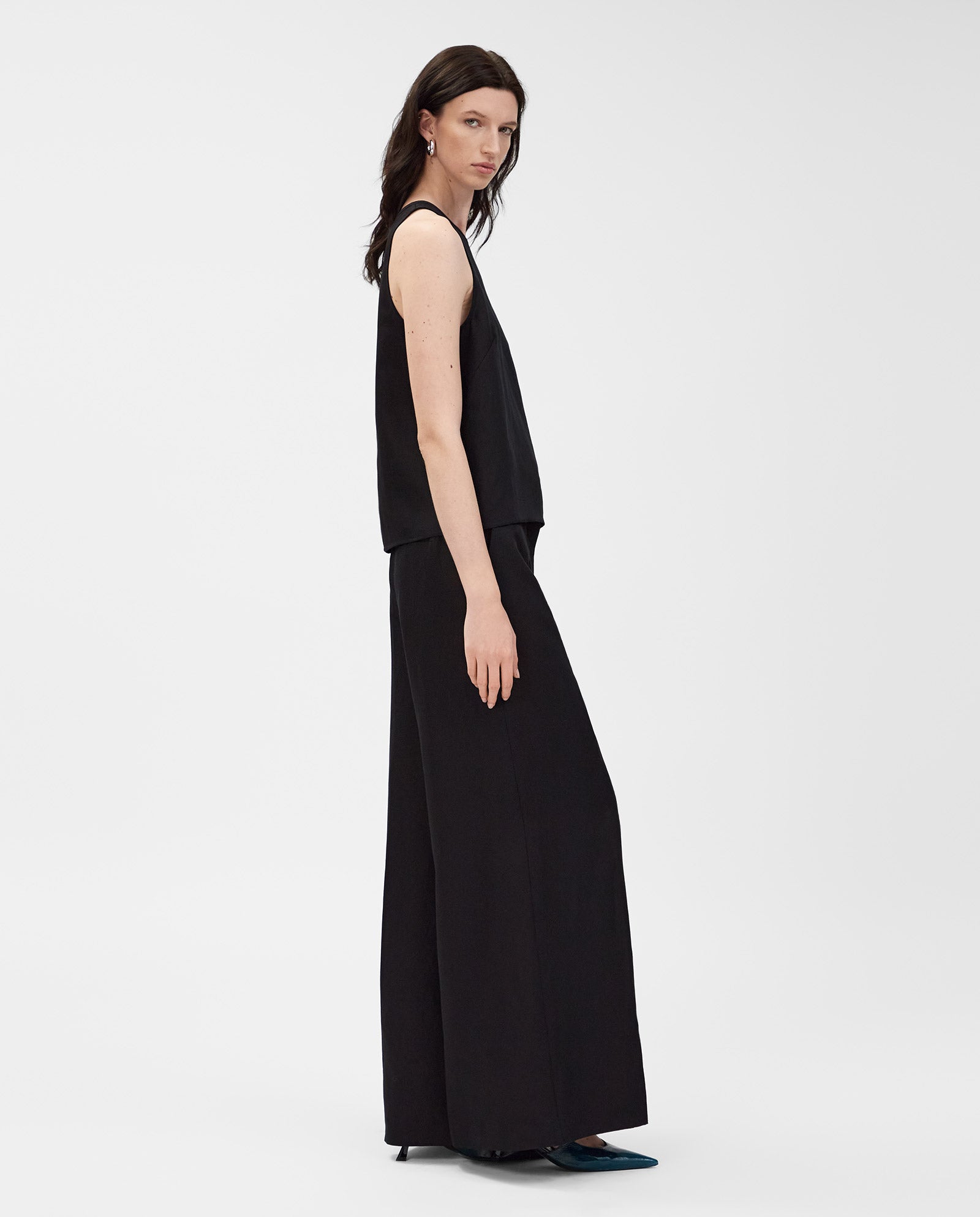 A woman stands sideways against a plain background, wearing the IVY OAK TAILOR SUE Top paired with wide-leg black linen-blend pants.