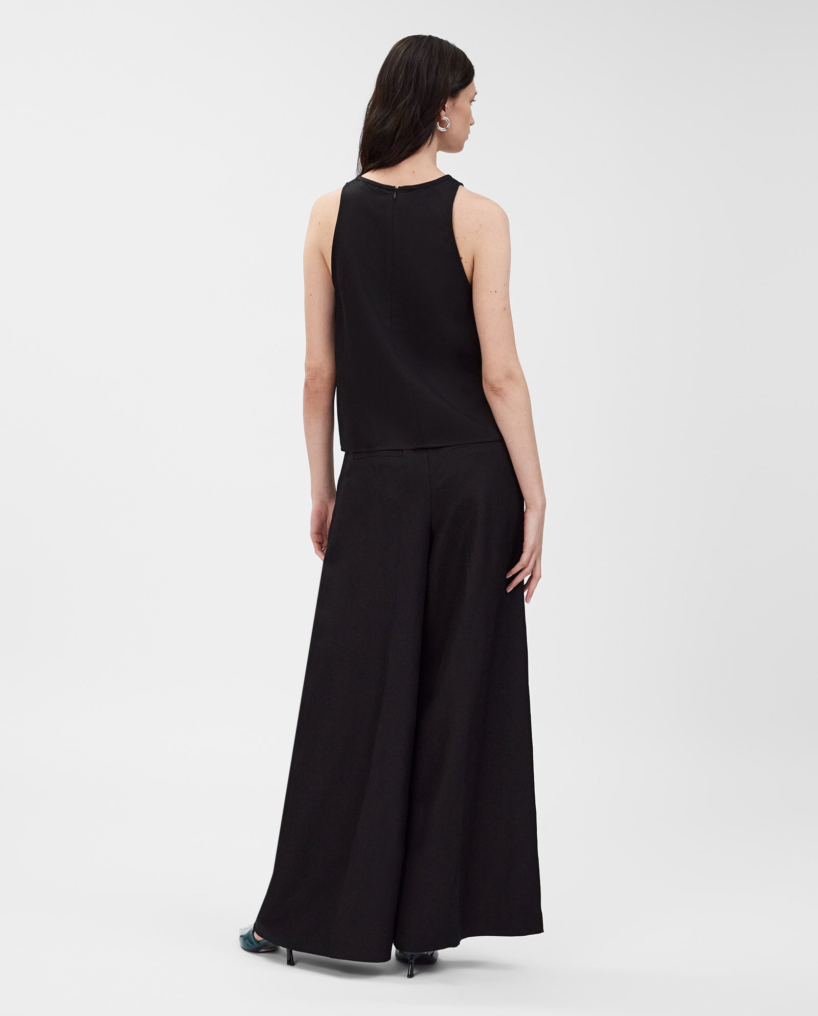 A woman stands with her back to the camera, wearing the IVY OAK TAILOR SUE sleeveless American shoulder top paired with wide-leg black linen-blend pants.