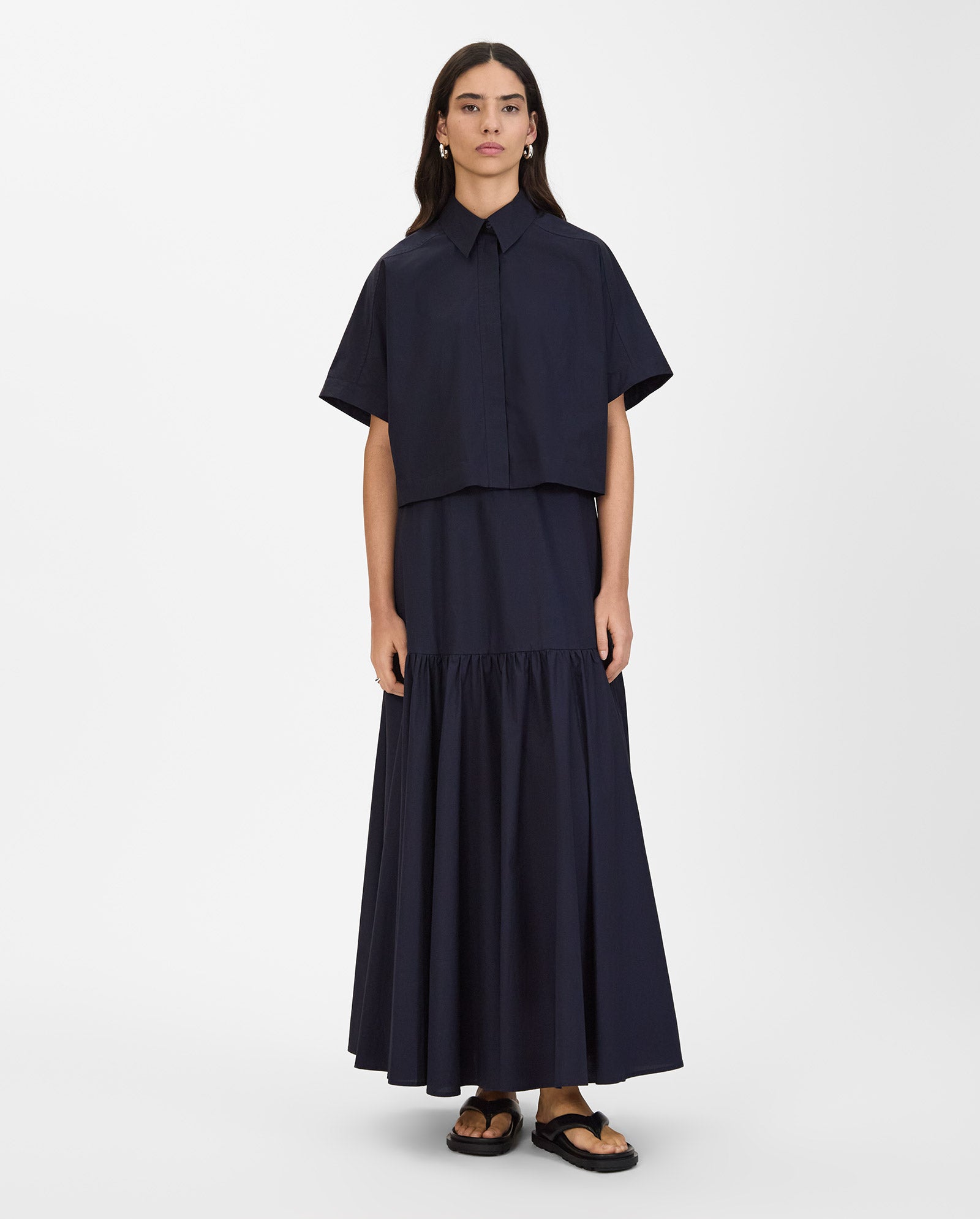 A woman wears the IVY OAK ELVIRA Shirt—a loose navy blouse with short, detachable sleeves—paired with a matching long skirt and black sandals.