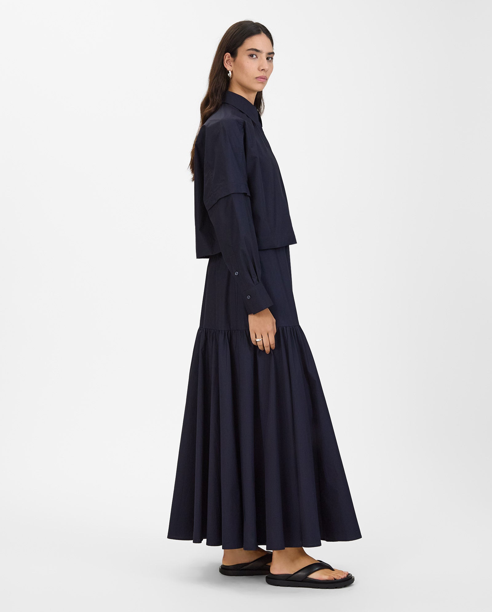 A woman stands sideways against a white background, wearing the IVY OAK ELVIRA Shirt with detachable sleeves, a long navy skirt, and black sandals.