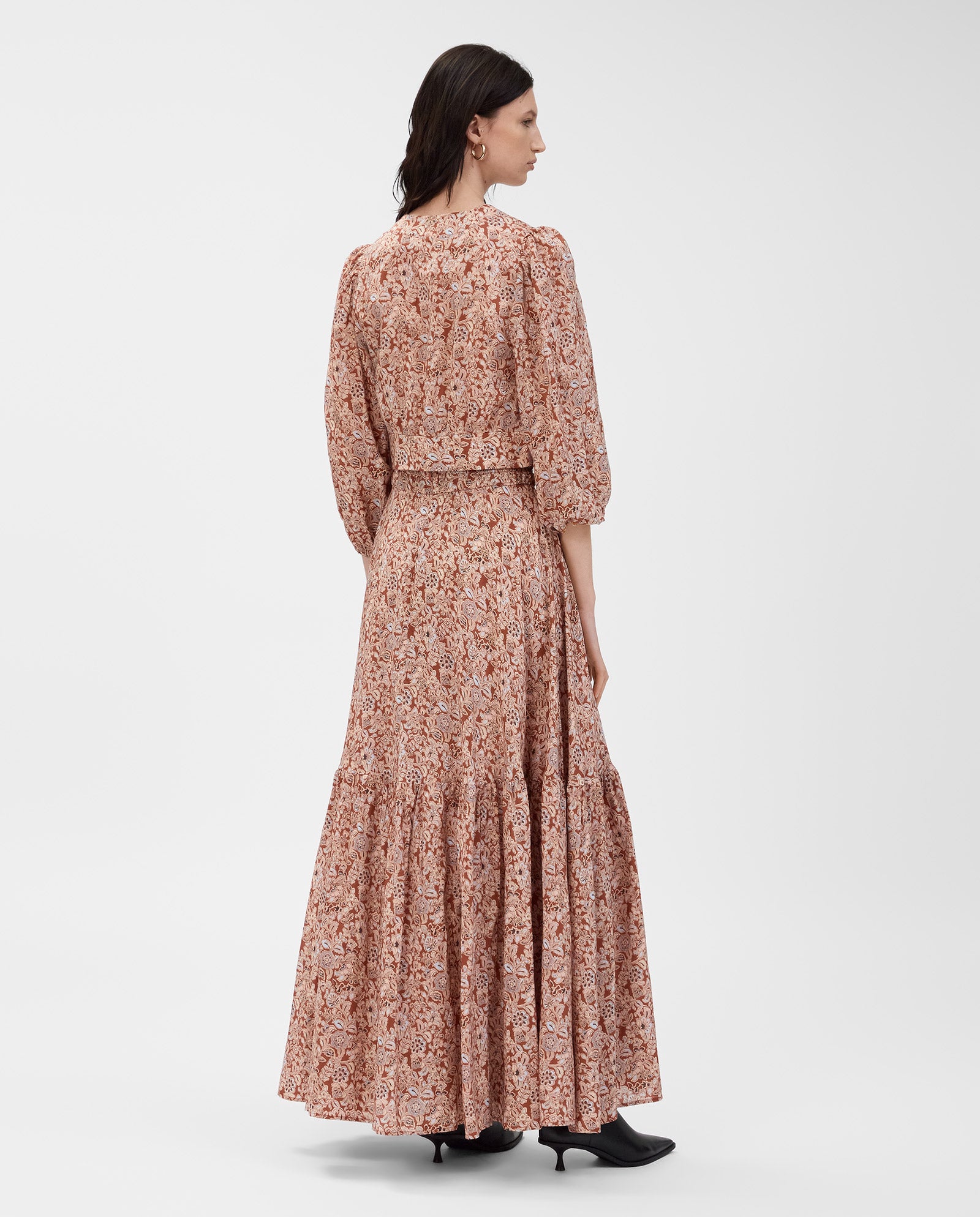 A woman stands facing away, wearing the IVY OAK ENRICA Blouse featuring a long, floral pink and brown design with a V-neck and balloon sleeves.