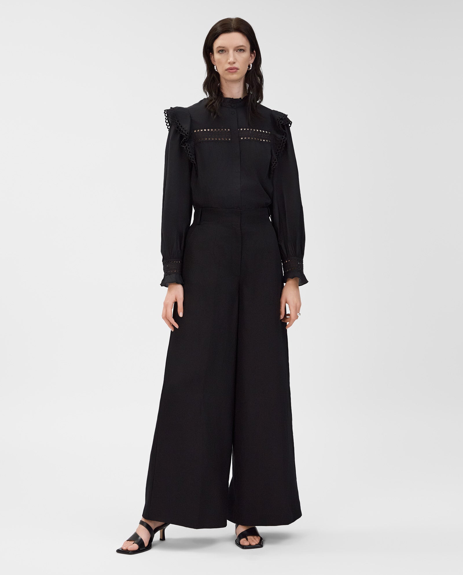 A woman stands against a plain background wearing IVY OAKs EVELINA Blouse—crafted from cotton voile with ruffled shoulders and lace tape—paired with black wide-leg pants.