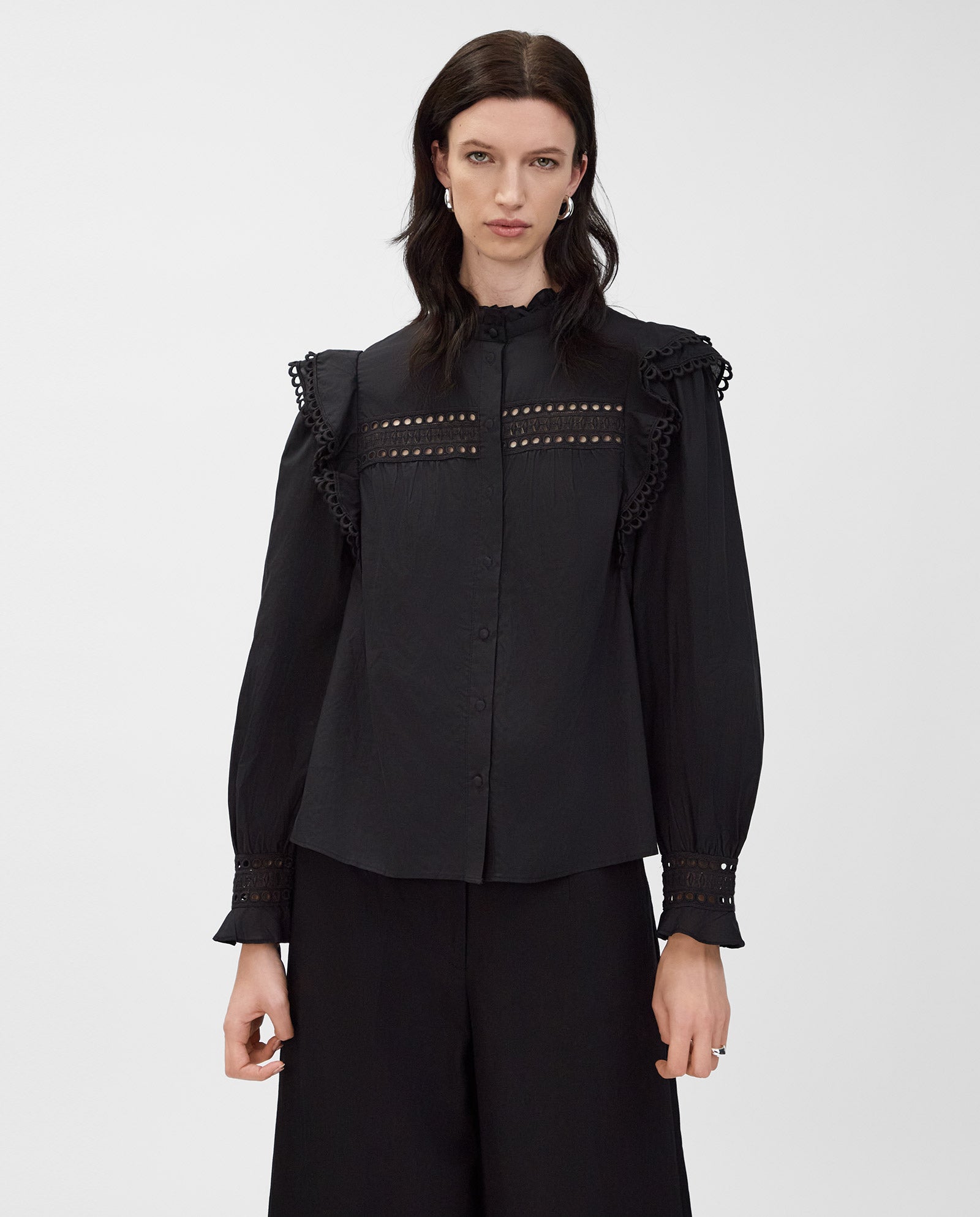 A woman wears the IVY OAK EVELINA Blouse in black cotton voile with lace tape details and wide-leg black pants, standing against a white background.