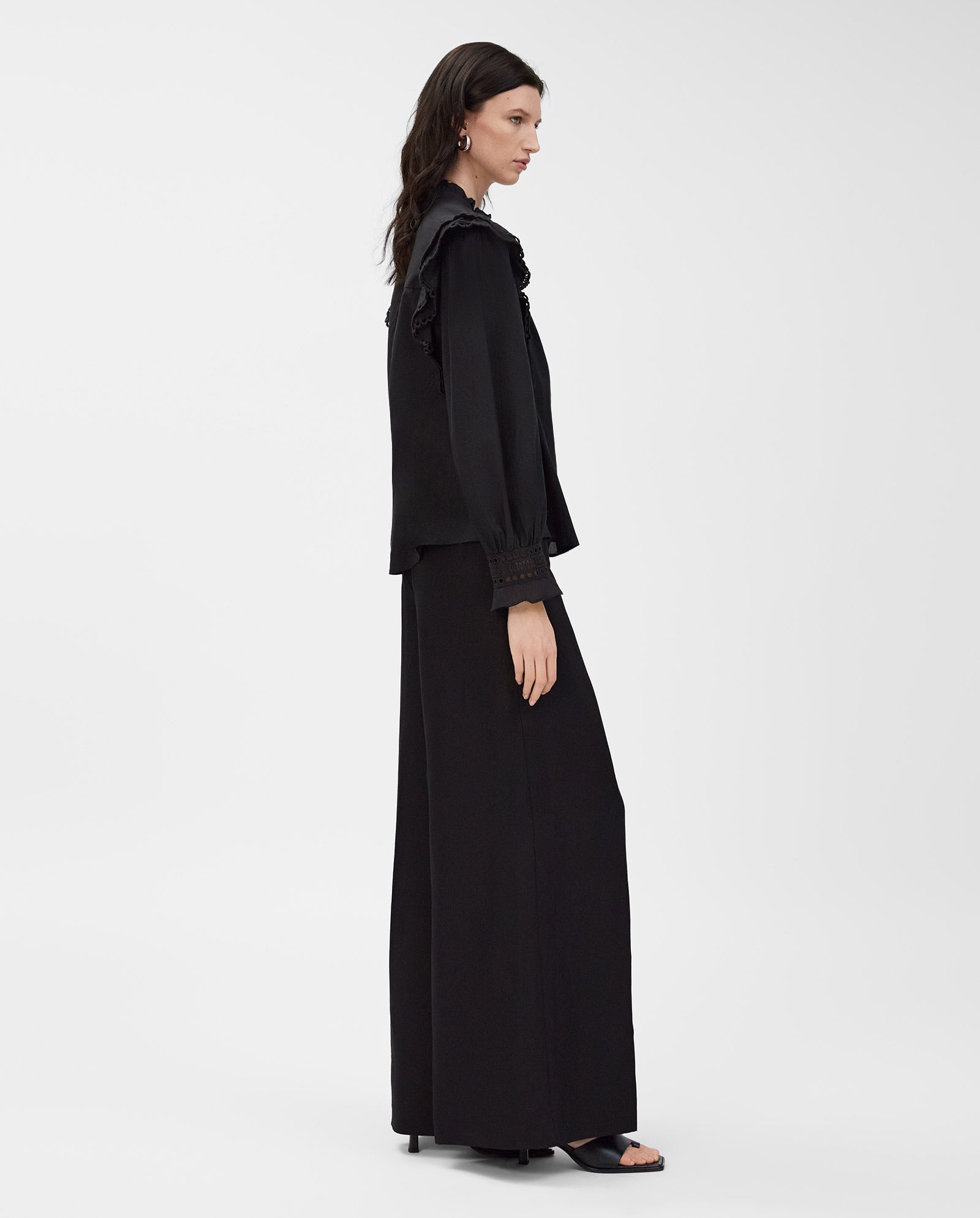 A woman in the IVY OAK EVELINA Blouse, made from cotton voile with a ruffled collar and lace tape, stands in profile against a white background, paired with wide-leg pants.