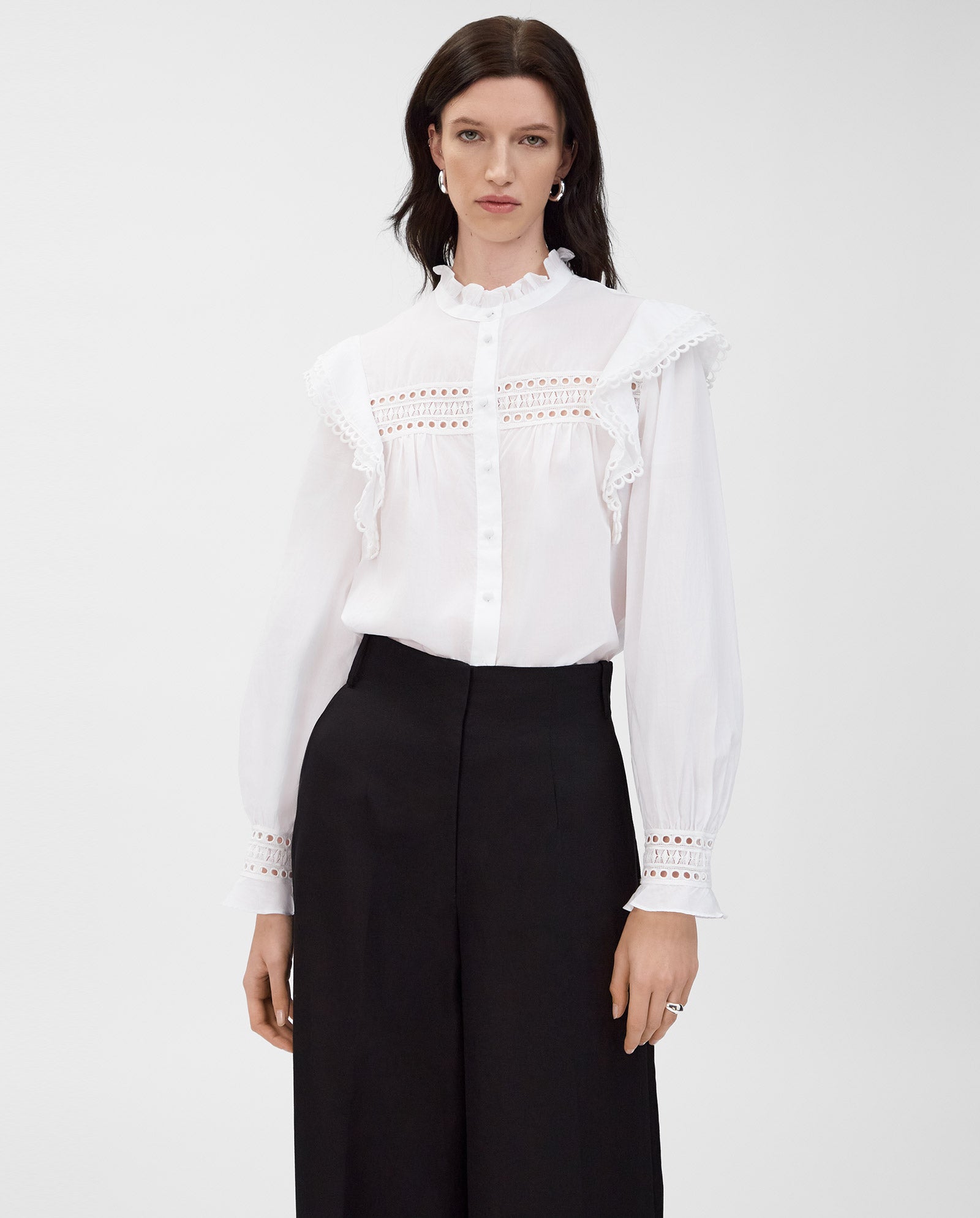 A woman wears the IVY OAK EVELINA Blouse in white cotton voile with lace tape details, paired with black wide-leg trousers, standing against a plain background.