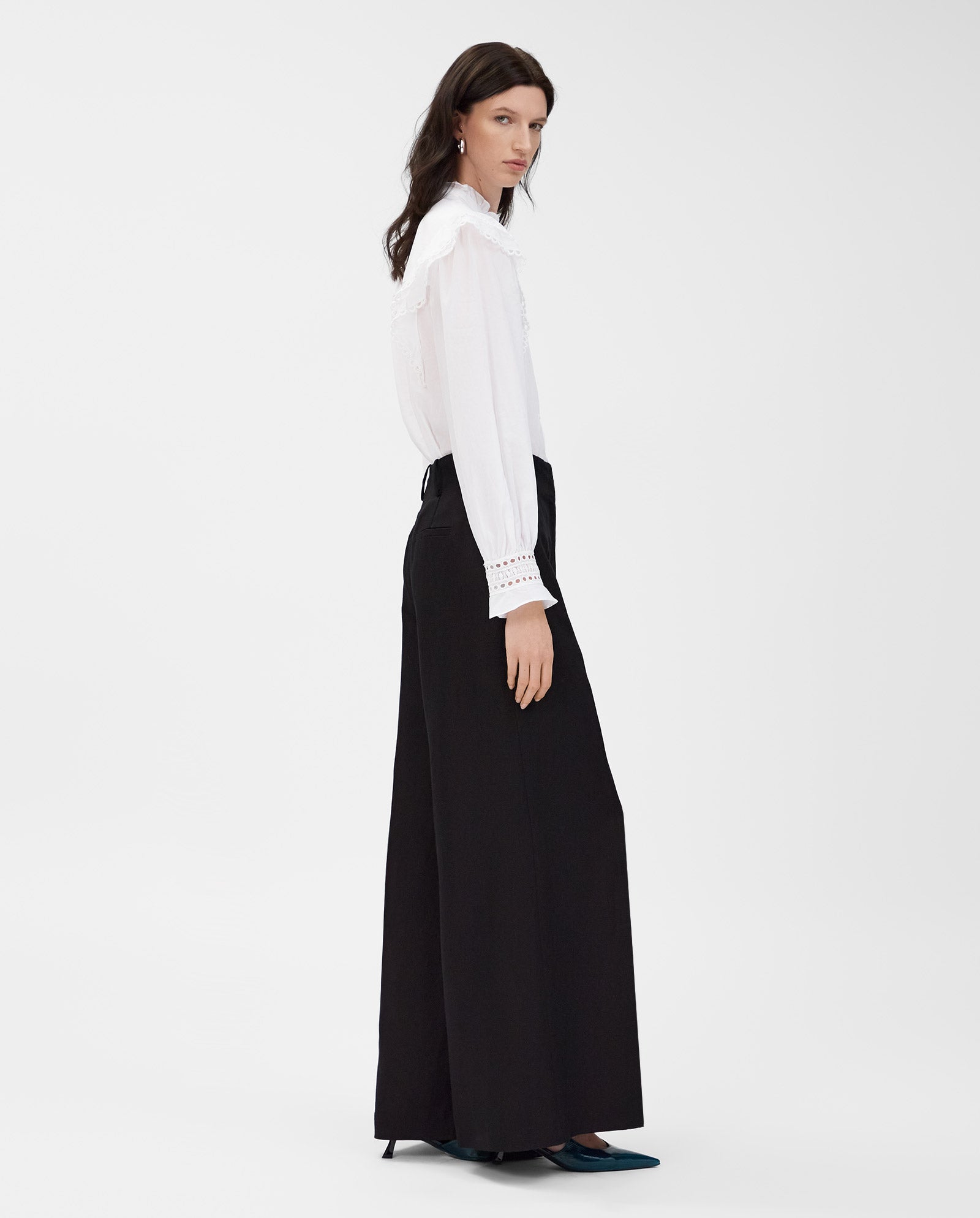 A woman wears the IVY OAK EVELINA Blouse in cotton voile with lace tape, paired with black wide-leg pants, standing sideways against a white background.