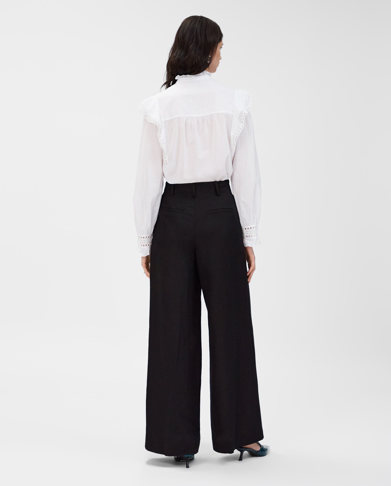 A woman faces away in the IVY OAK EVELINA Blouse, crafted from white cotton voile with delicate lace tape, paired with black wide-leg pants against a white background.