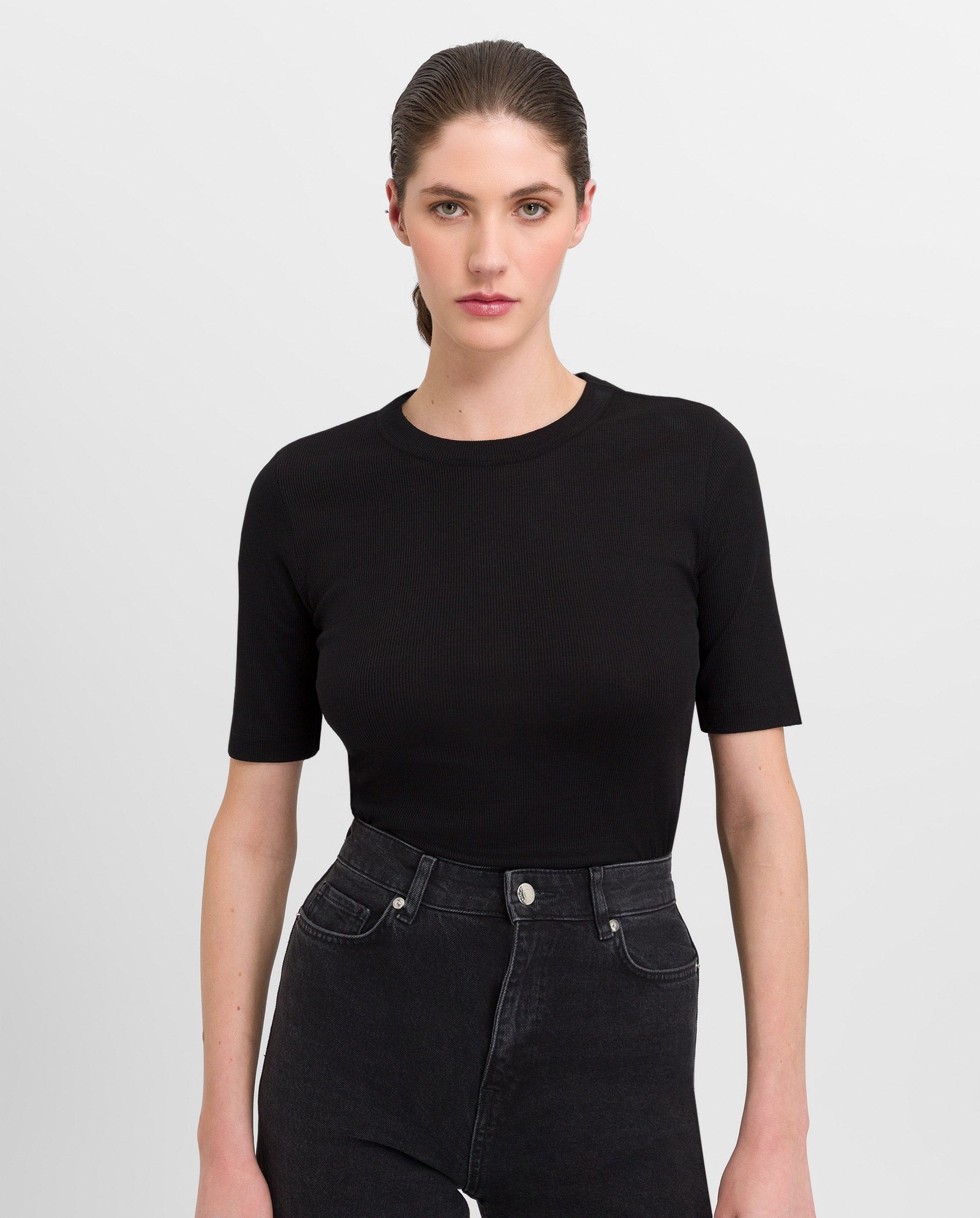 A woman with straight brown hair, wearing the IVY OAK KRISTIN Rib T-Shirt in black and high-waisted black jeans, stands facing forward.