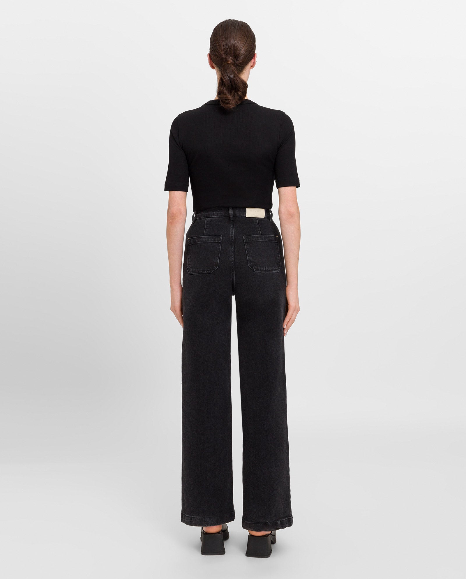 A woman stands facing away, wearing the IVY OAK KRISTIN Rib T-Shirt in black, paired with high-waisted black wide-leg jeans and heeled shoes.