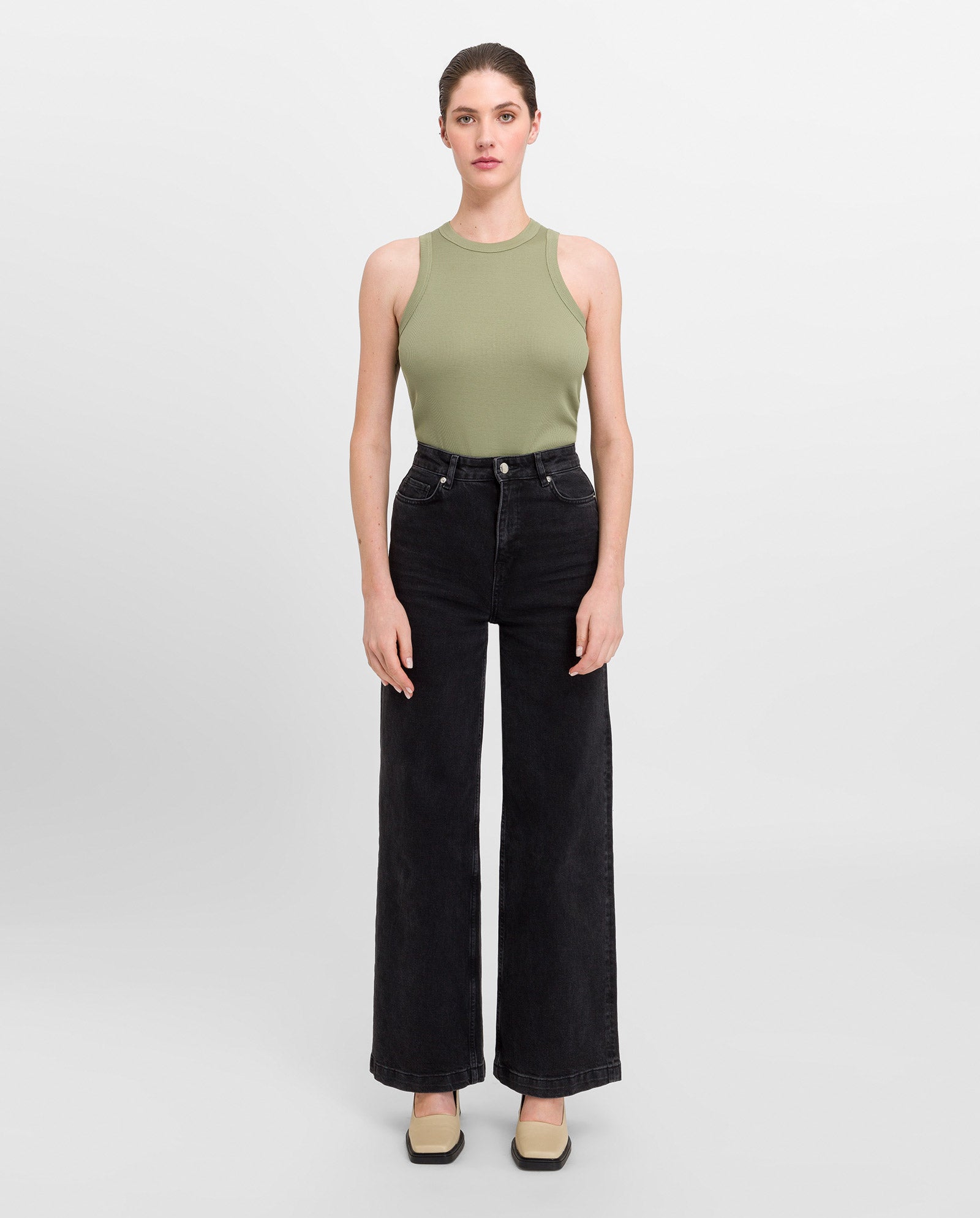 A woman wears the IVY OAK KLARA ANN sleeveless fitted green top made from organic cotton, paired with black wide-leg jeans, standing against a plain light background.