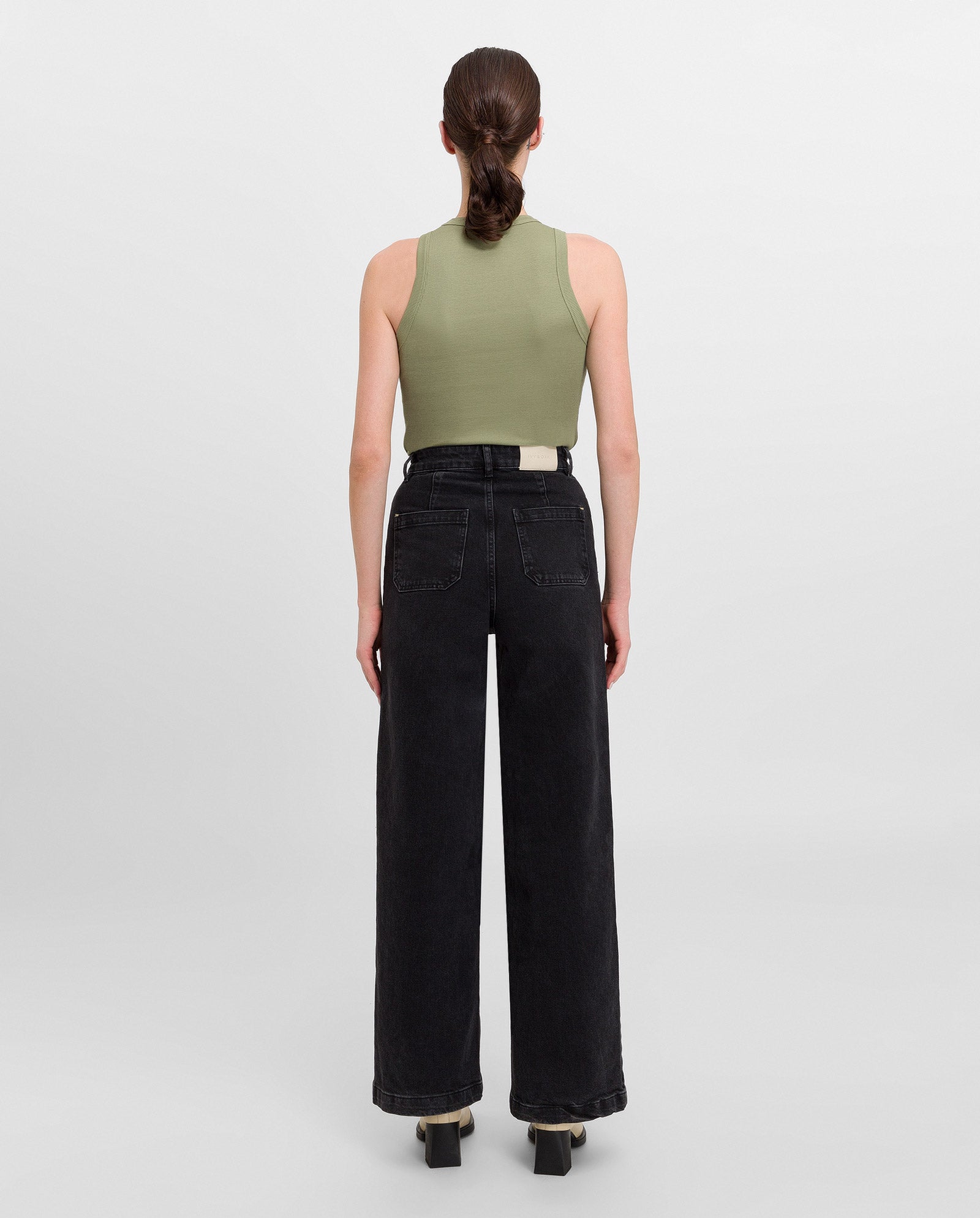 A woman faces away, wearing the IVY OAK KLARA ANN Sleeveless Top in olive green organic cotton jersey and black wide-leg jeans, standing against a plain background.