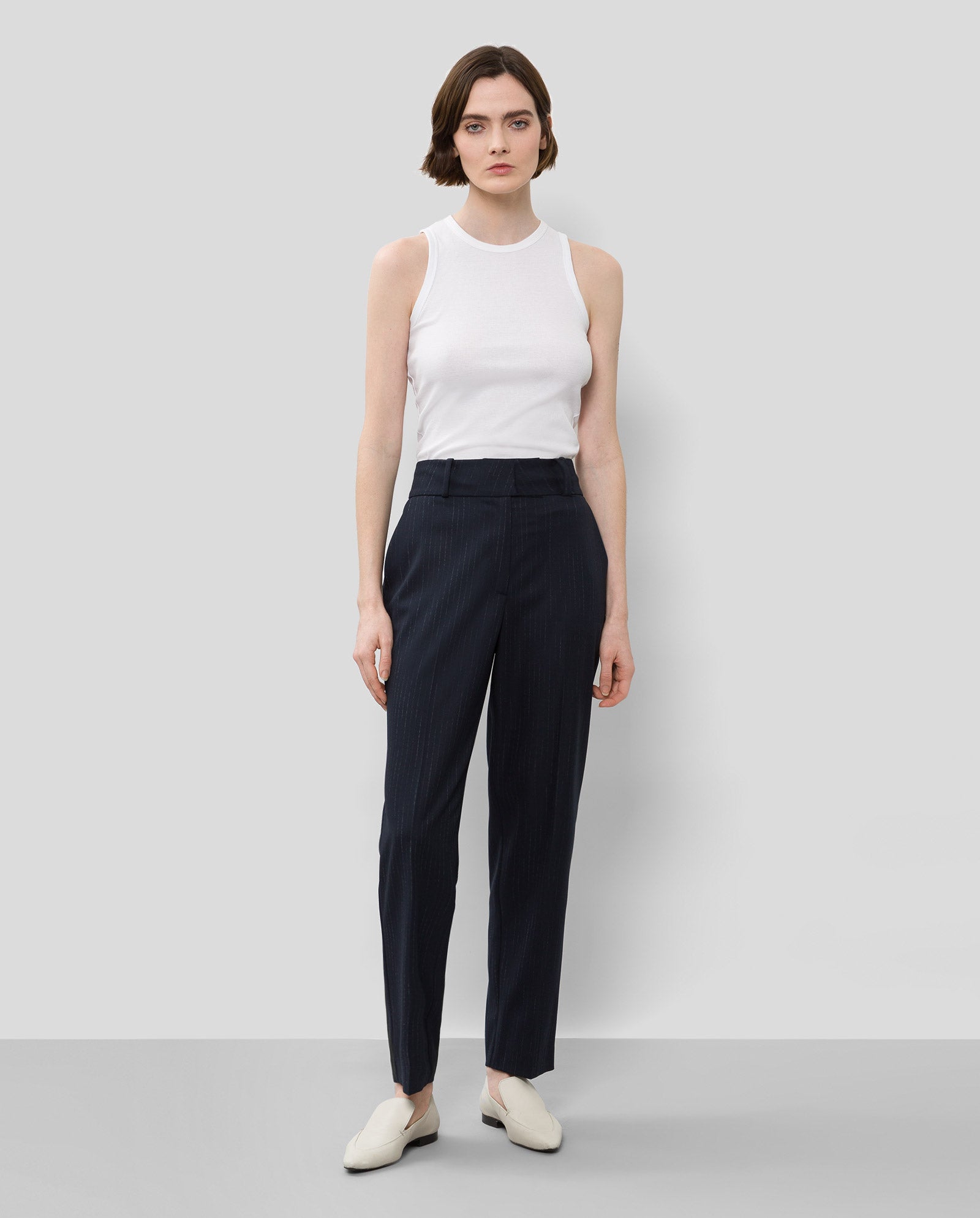 A woman stands against a light background wearing a white sleeveless top, white shoes, and IVY OAKs PHOEBE ANN Trouser—high waist pinstripe pants crafted from recycled polyester.