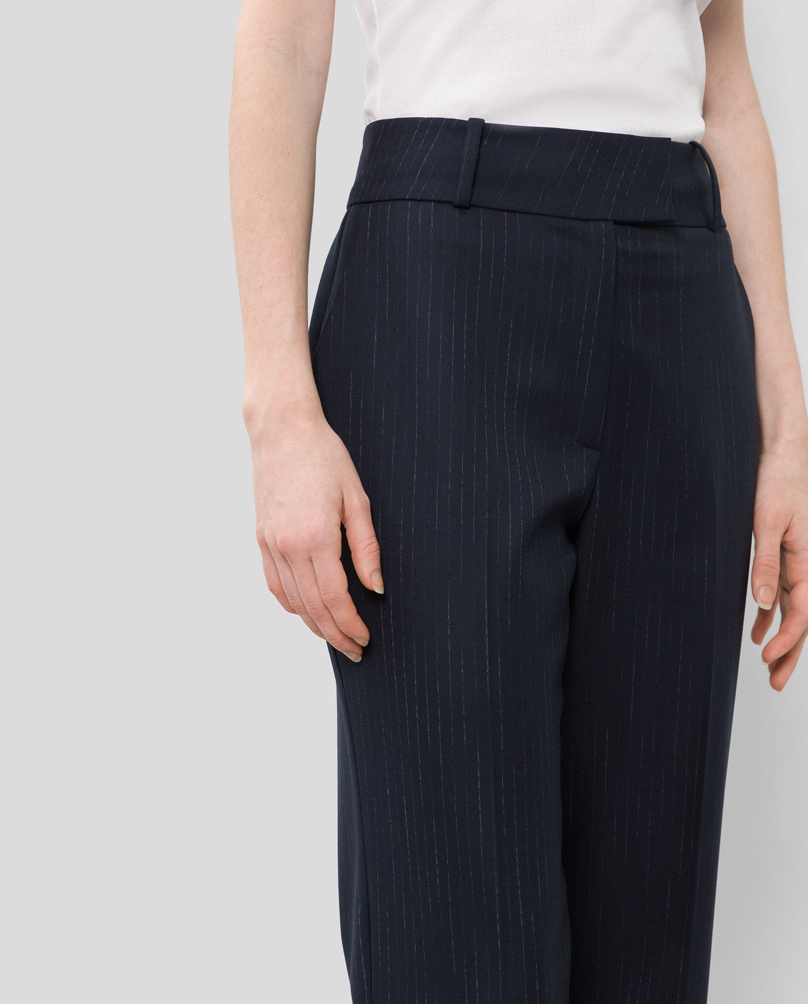 A model wears the IVY OAK PHOEBE ANN Trouser, featuring a high waist and pinstripes, crafted from recycled polyester, paired with a white top. The outfit is shown from shoulder to knee against a plain background.