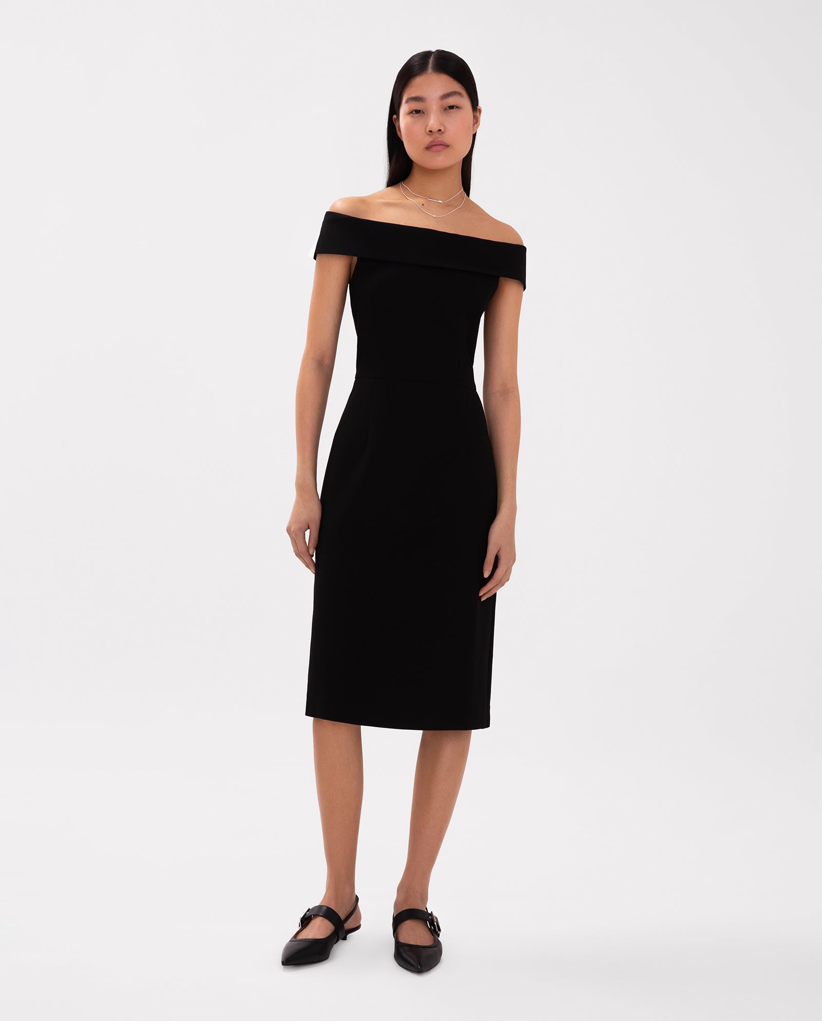 A woman wearing the IVY OAK MAREN fitted black off-shoulder dress with black flats stands against a plain white background.
