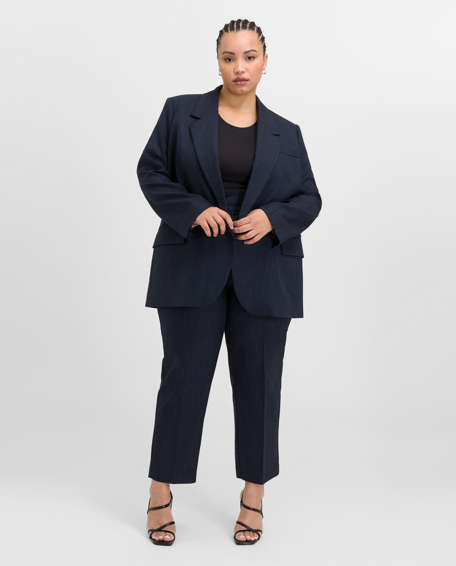 A woman faces forward against a plain white background, wearing the IVY OAK JANINA ANN Blazer with piped pockets and pinstripes, paired with black heels.