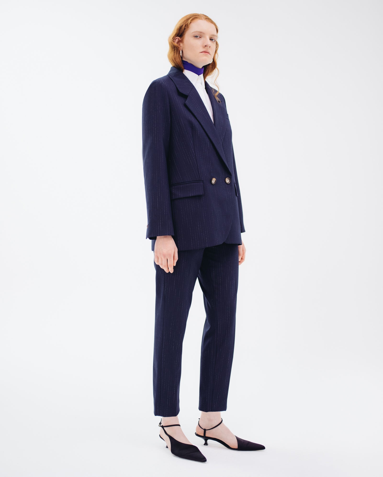 Someone wearing the IVY OAK JANINA ANN Blazer, a navy pinstripe design with piped pockets, paired with a white shirt and black heels, stands against a plain white background.