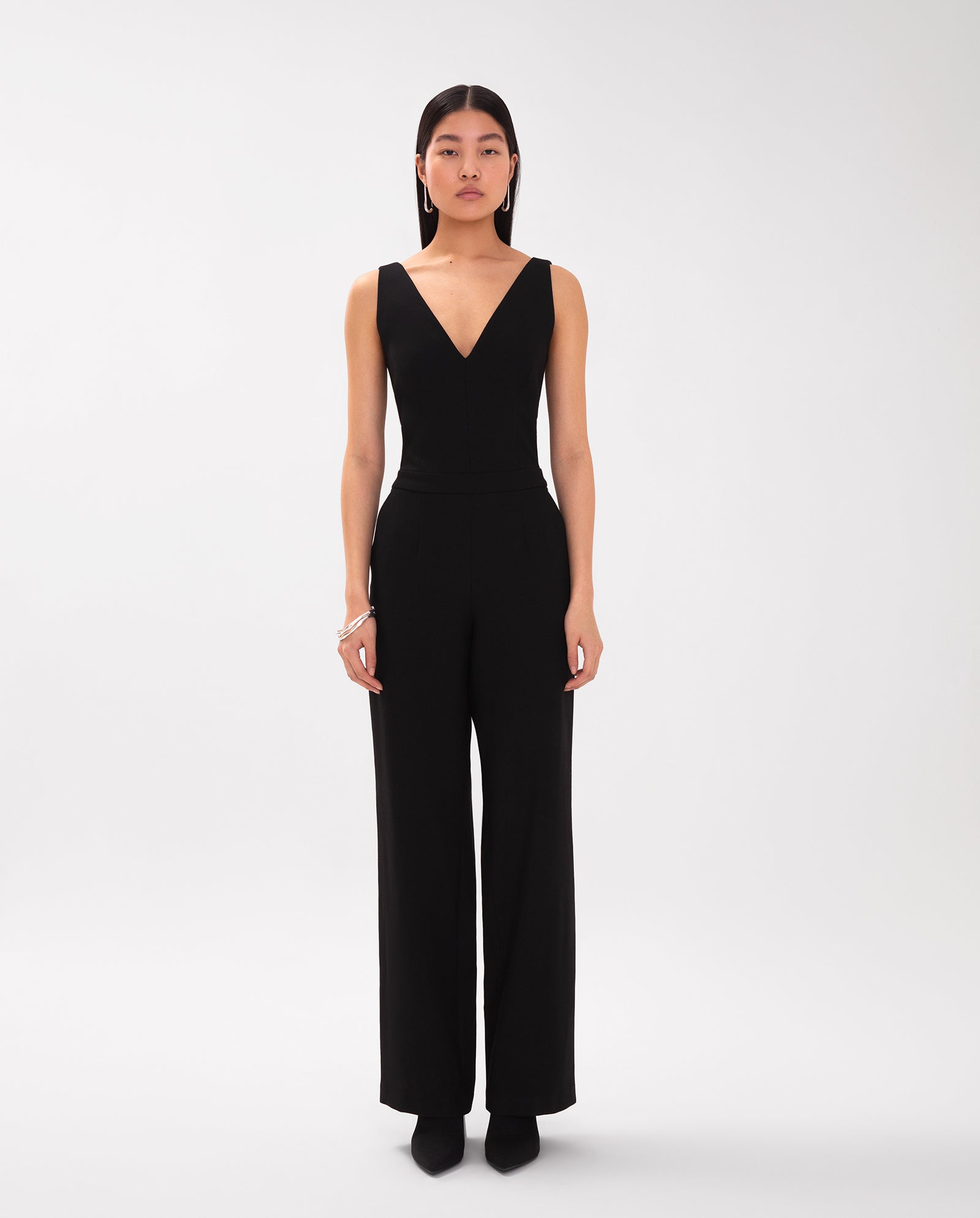A woman stands against a white background wearing the IVY OAK PAULINA Jumpsuit, featuring a sleeveless black design with a V-neck, wide legs, and a tailored waistband.