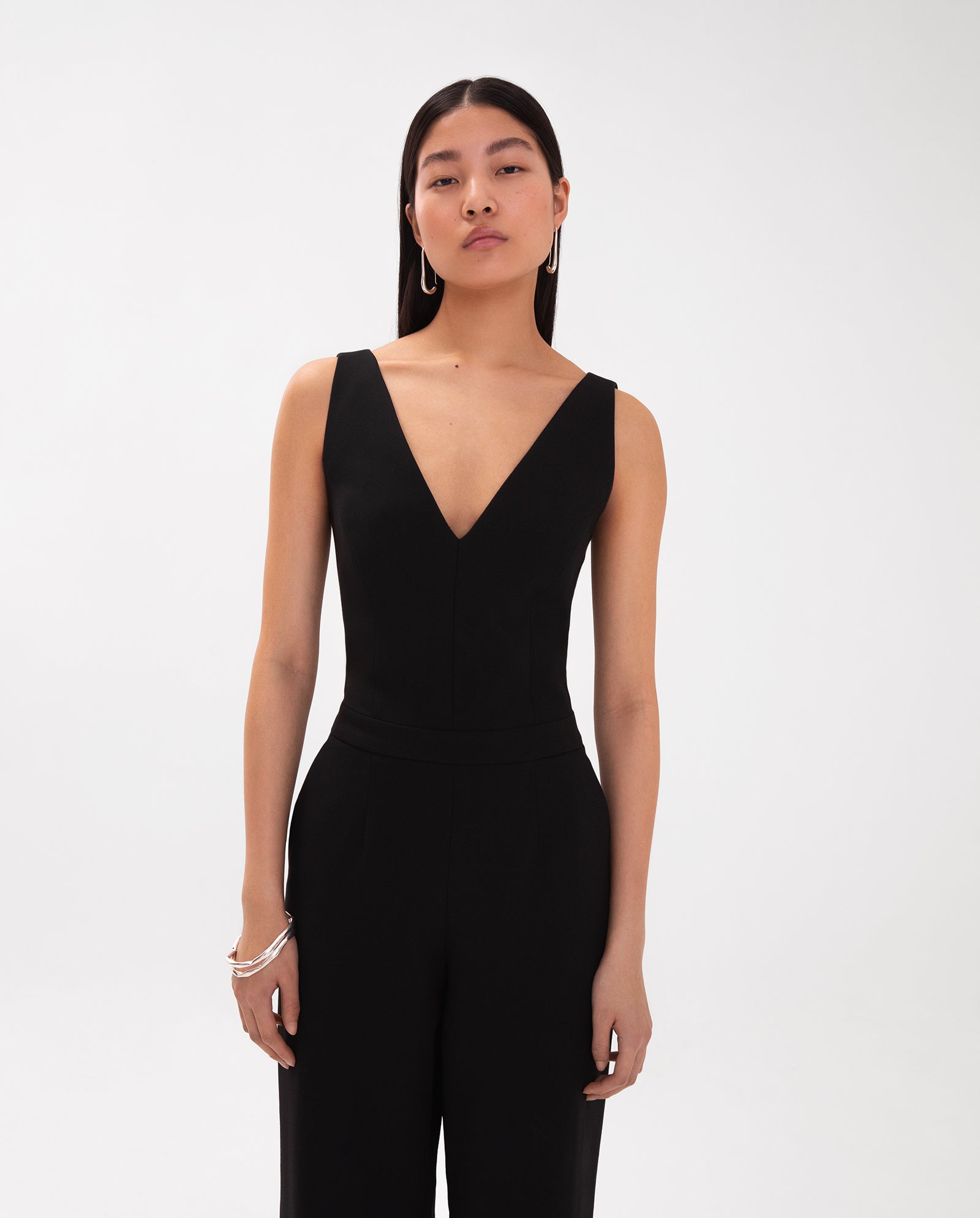 A woman wears the IVY OAK PAULINA Jumpsuit—a sleeveless black V-neck style with a defined waistband—standing against a plain white background.