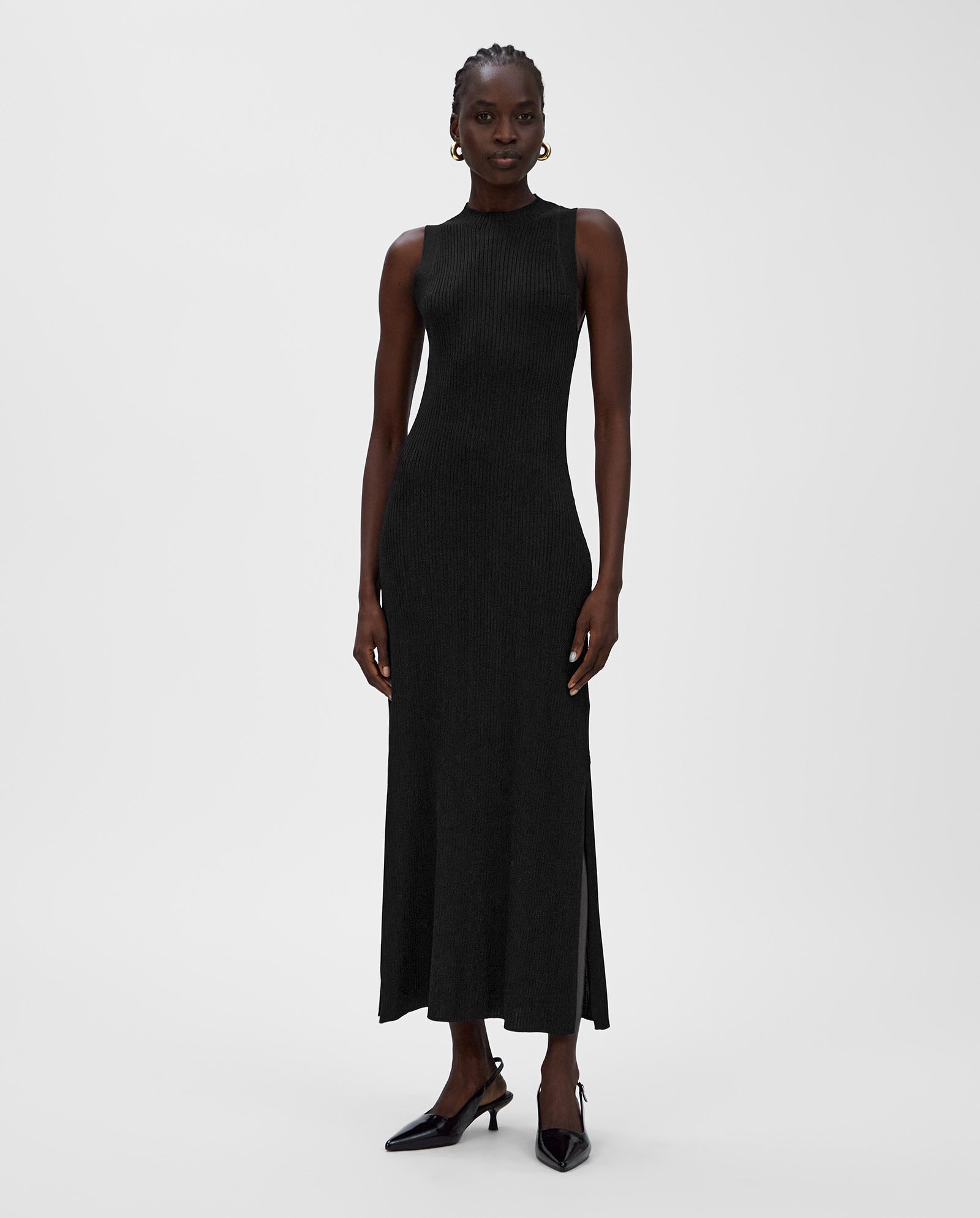 A woman in a sleeveless, black KLAUDIA Dress by IVY OAK with rib structure and black heels stands against a plain white background.