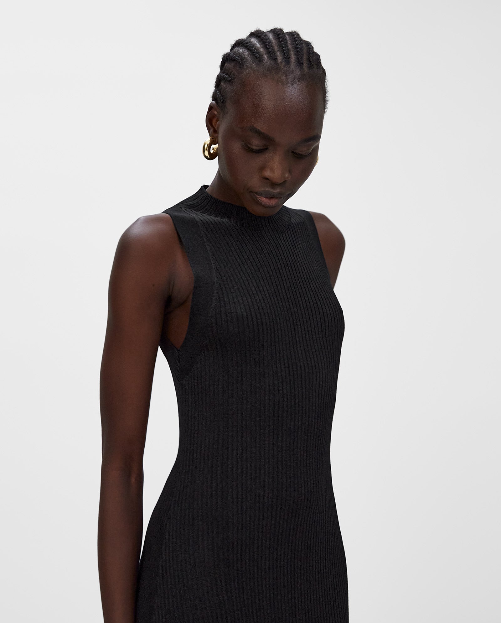 A woman wears the sleeveless black KLAUDIA Dress by IVY OAK, featuring a distinctive rib structure, as she looks down against a plain white background.